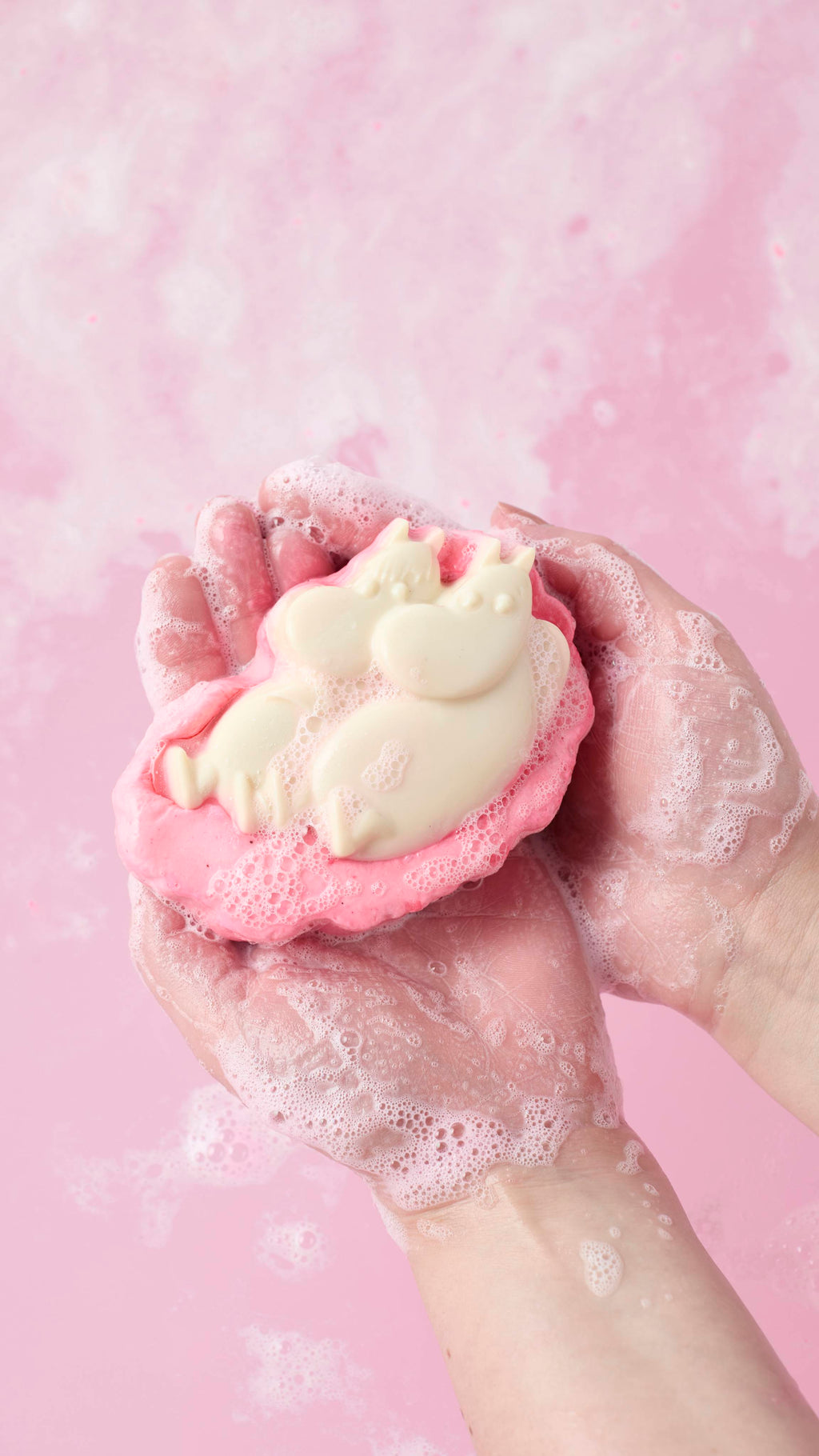 An overhead shot of two soapy hands cradling a pink and cream-coloured "Moomin Delight" soap and body wash product. White foam covers the skin, set against a background of swirling, bubbly pink water.