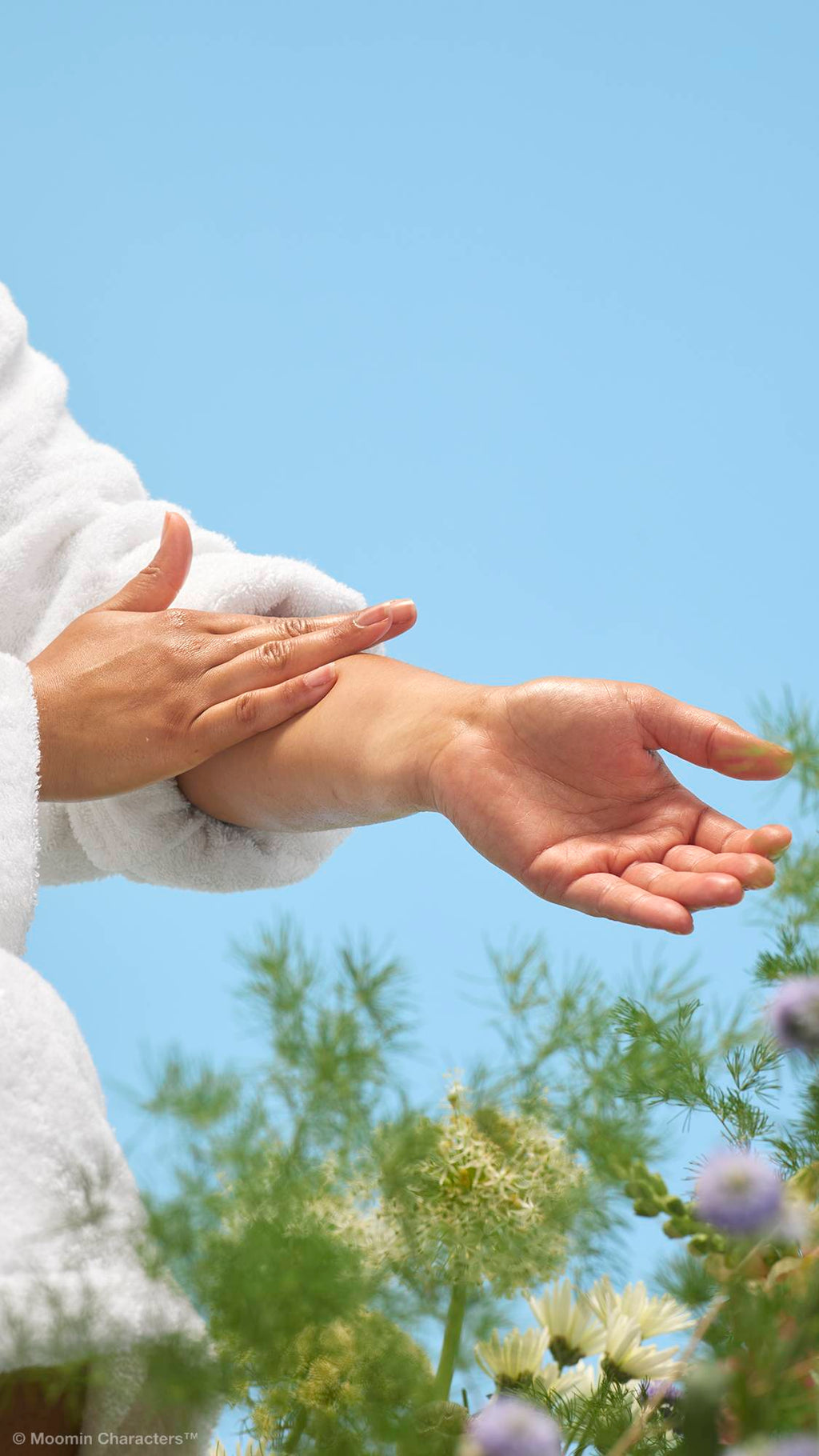 A close-up shot of a person in a white dressing gown smoothing the cream-coloured "Moominmamma" lotion onto their forearms and hands. Soft green foliage and a white flower are visible in the background against a clear blue sky.