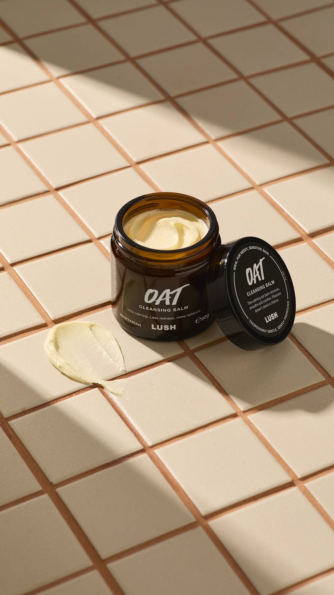 An open brown glass jar of Lush "Oat" facial cleanser sits on a beige tiled surface. A thick dab of the pale yellow, creamy product is smeared on the tiles next to the jar and its black lid.