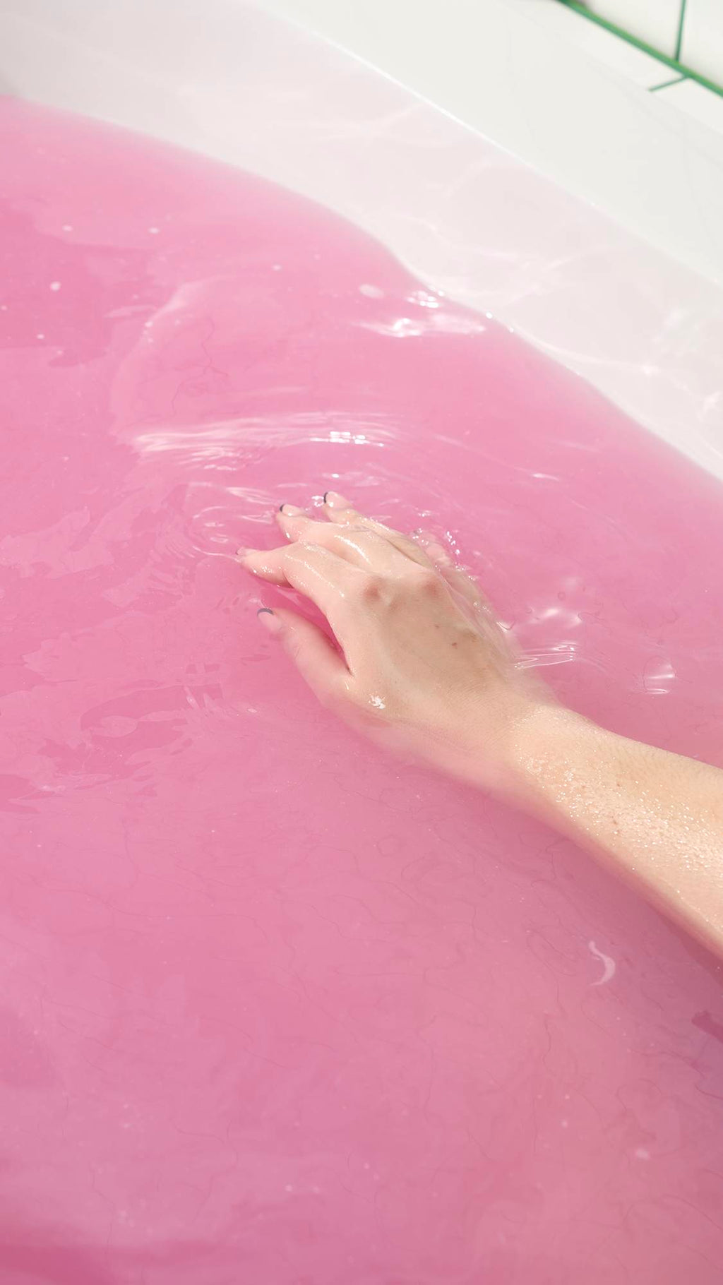 The Sex Bomb Bath Bomb has completely dissolved, leaving the bathtub filled with a vibrant, opaque shade of deep pink. A hand is shown gently submerged in the colorful water, illustrating the relaxing and aesthetically pleasing result of the bath.