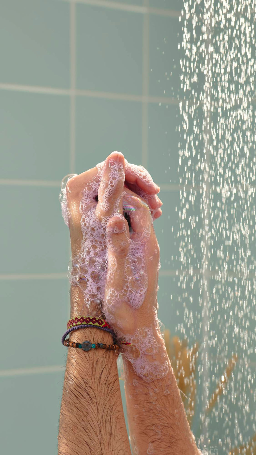 A close-up image of a model's hands covered in thick, pale purple "Stormy Weather" shower gel as they lather in the shower. A stream of falling water is visible on the right against light green tiles.