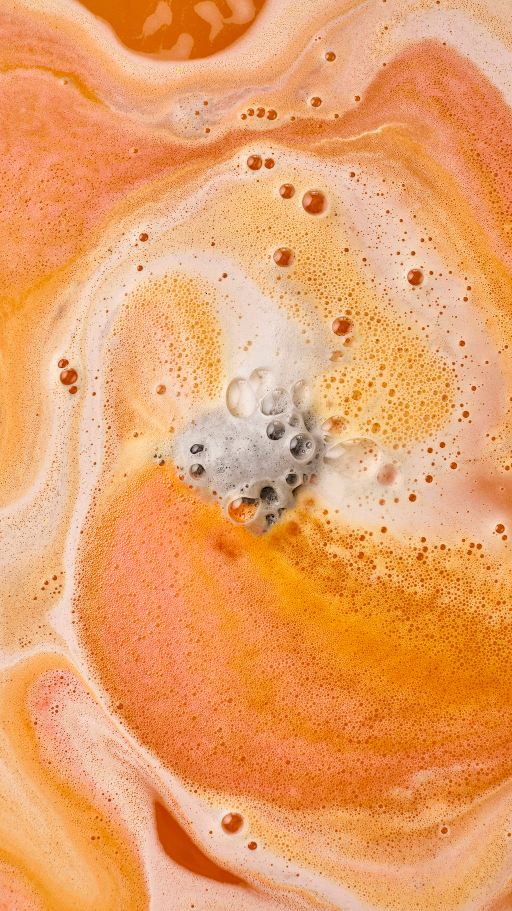 An aerial view of orange and yellow water swirling with thick, cream-coloured foam. In the centre, a grey "Volcano" foot soak product dissolves, releasing small bubbles and creating an orange and white marbled effect across the surface.