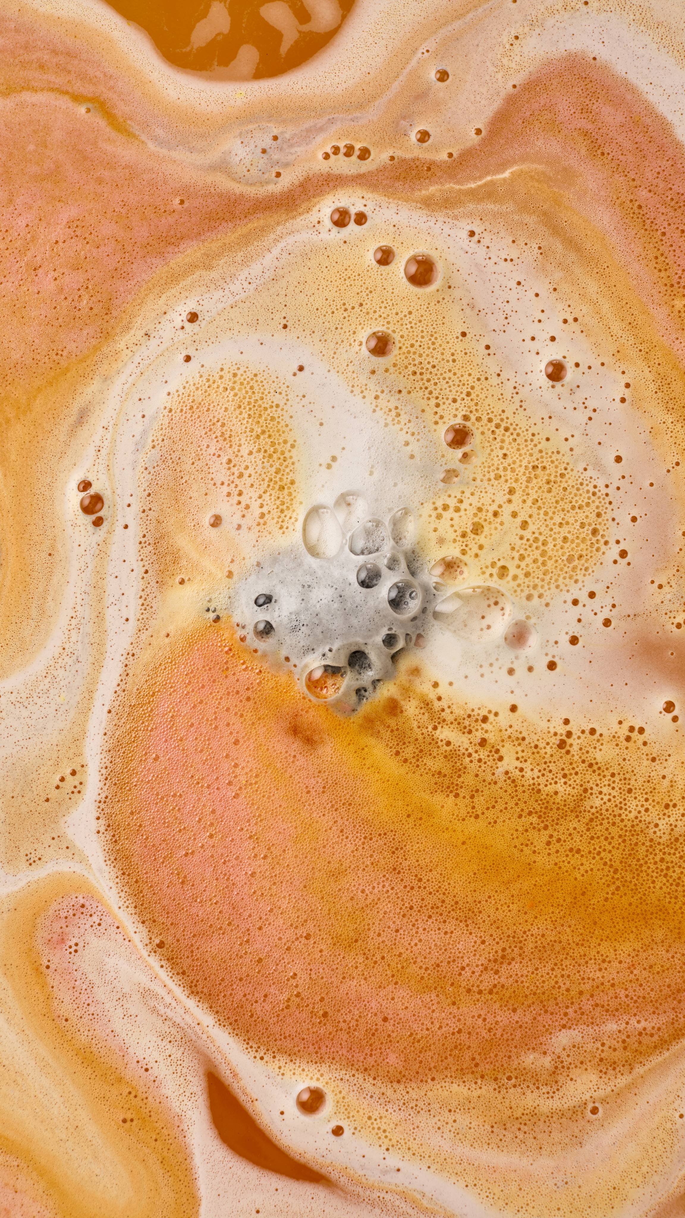 An aerial view of orange and yellow water swirling with thick, cream-coloured foam. In the centre, a grey "Volcano" foot soak product dissolves, releasing small bubbles and creating an orange and white marbled effect across the surface.