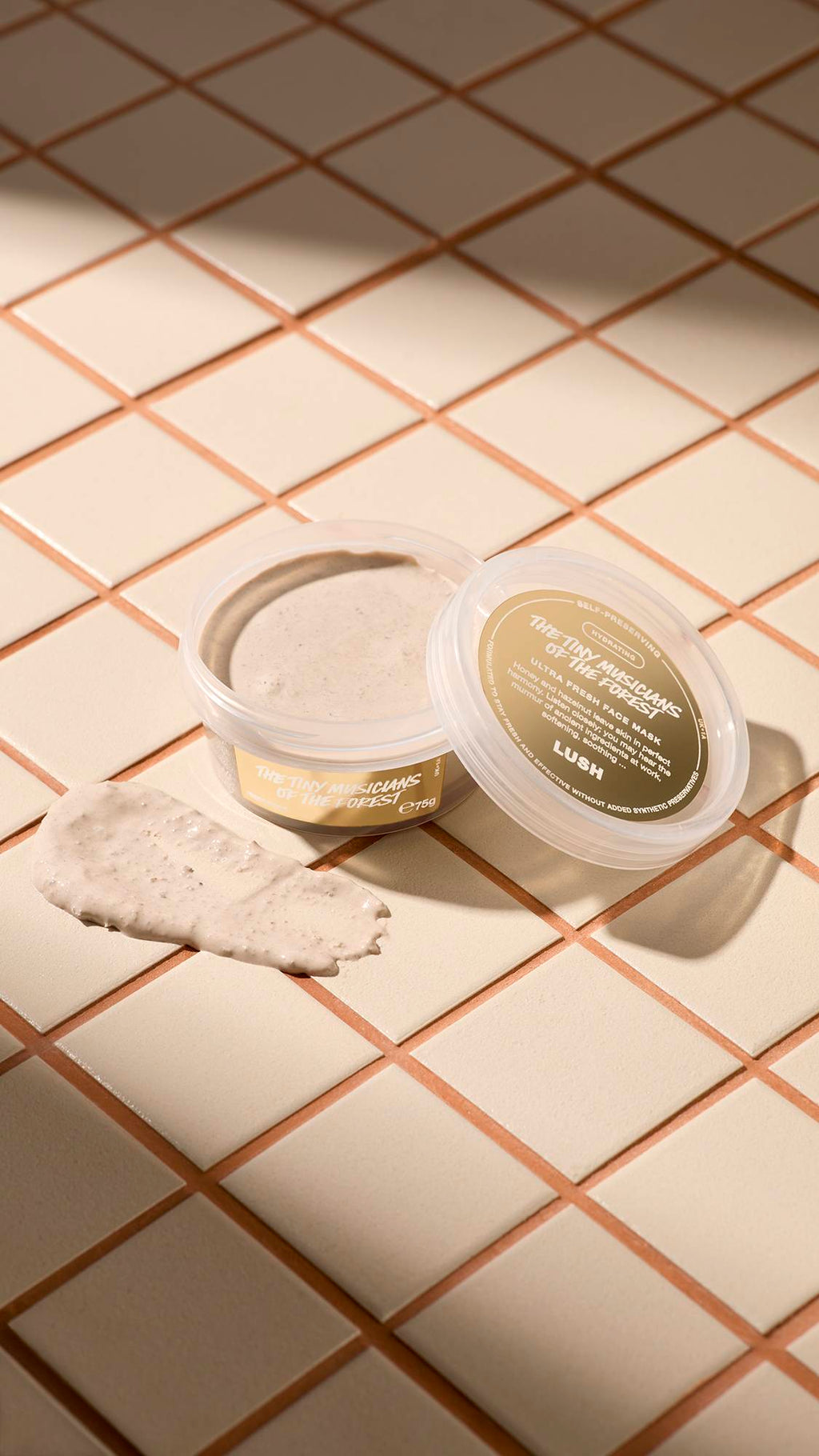 An open, clear tub of Lush "The Tiny Musicians Of The Forest" face mask sits on light beige tiles next to its lid. A thick dollop of the grainy, beige-coloured mask is smeared on the tiles in front of the pot.