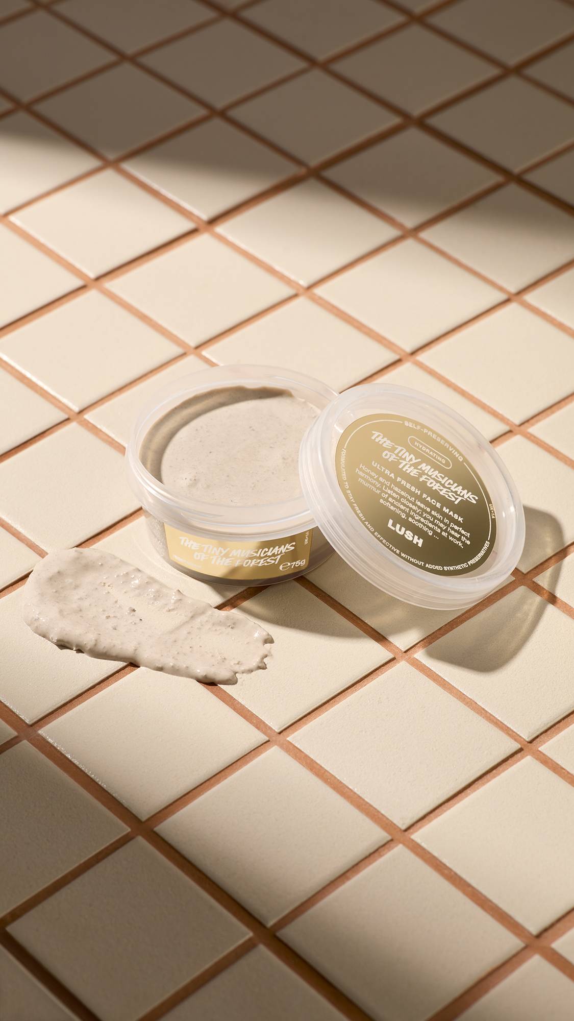 An open, clear tub of Lush "The Tiny Musicians Of The Forest" face mask sits on light beige tiles next to its lid. A thick dollop of the grainy, beige-coloured mask is smeared on the tiles in front of the pot.
