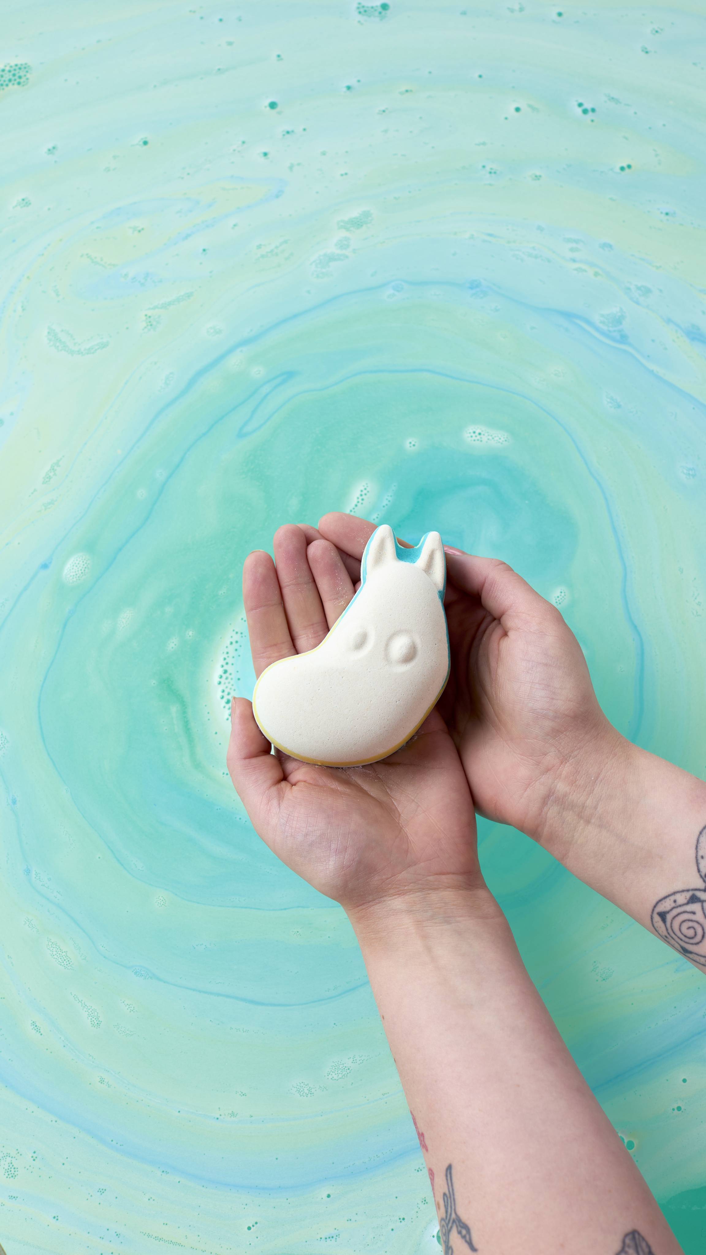 An overhead shot of two hands holding a white Moomintroll bath bomb over swirling turquoise and light green bathwater. The water creates a circular, marbled pattern around the hands, which have tattoos visible on the forearms.