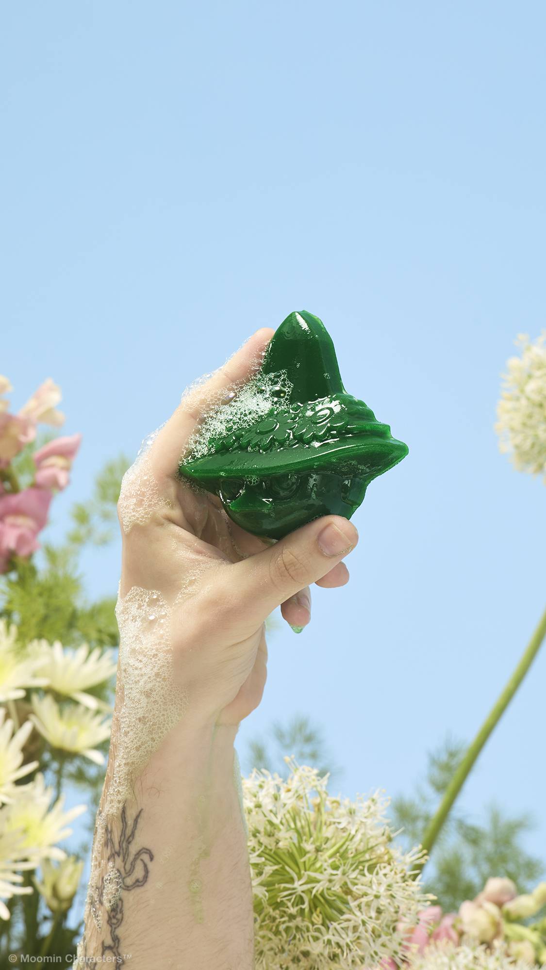 A close-up shot of a tattooed arm holding up a green, soapy Snufkin-shaped shower gummy covered in frothy bubbles. Yellow, white, and pink wildflowers are visible in the background against a clear blue sky.