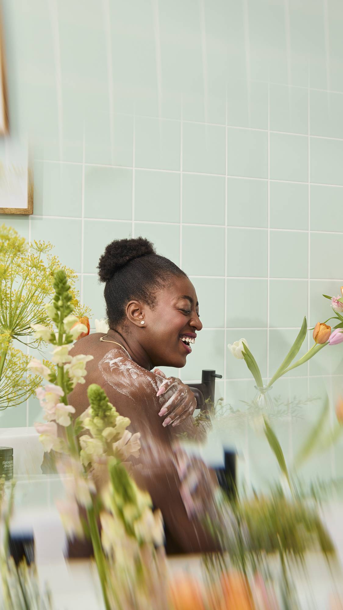 A smiling woman with dark hair in a bun lathers her shoulder with soapy suds while sitting in a bathtub. The joyful scene is framed by blurred yellow and pink flowers against a mint green tiled wall.