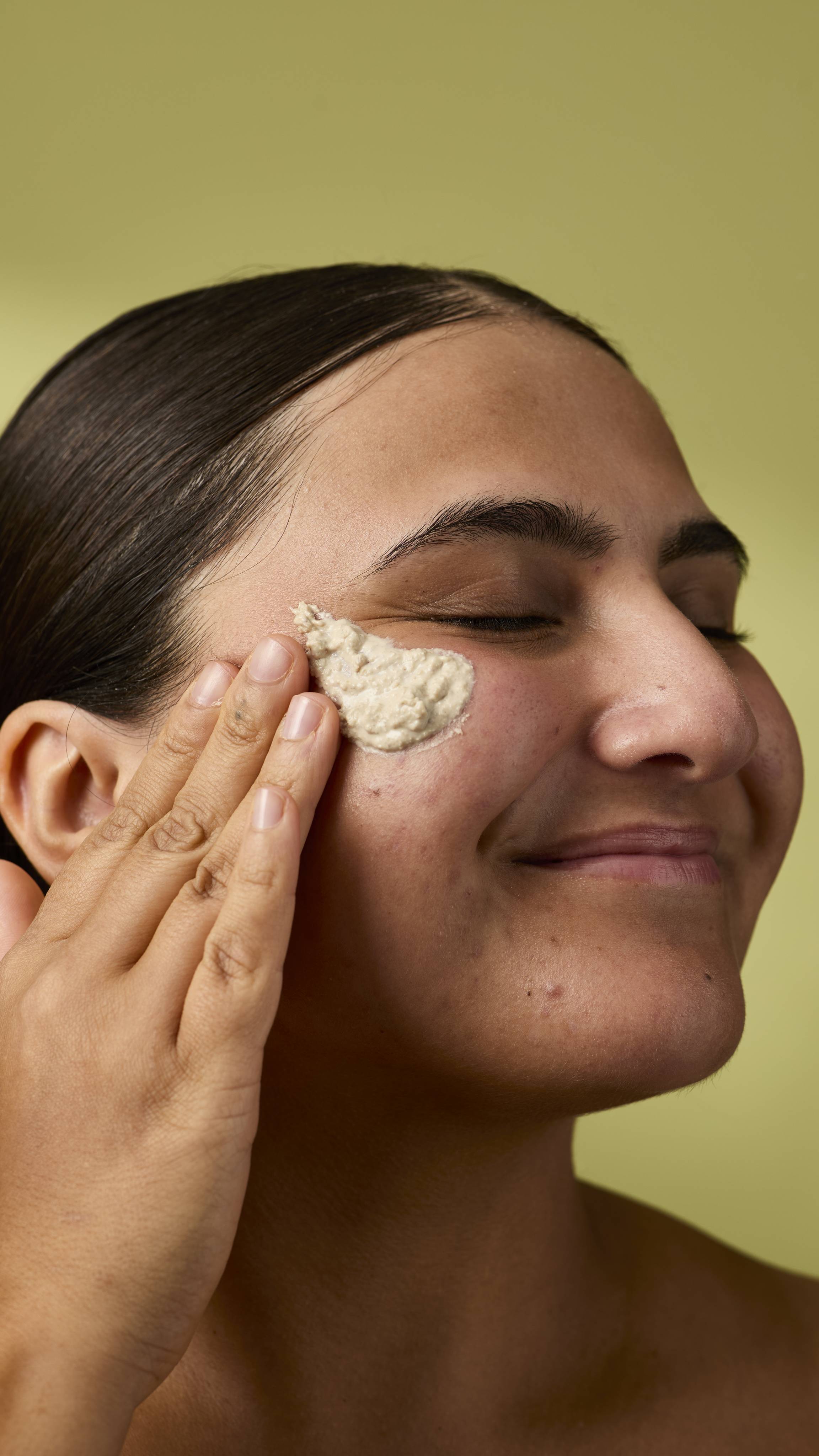 A close-up image of the model's face on an olive-green background. Their dark hair is tied back, and they are applying the textured Biomic facial cleanser to the skin on their cheek with their eyes closed.