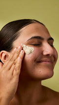 A close-up image of the model's face on an olive-green background. Their dark hair is tied back, and they are applying the textured Biomic facial cleanser to the skin on their cheek with their eyes closed.
