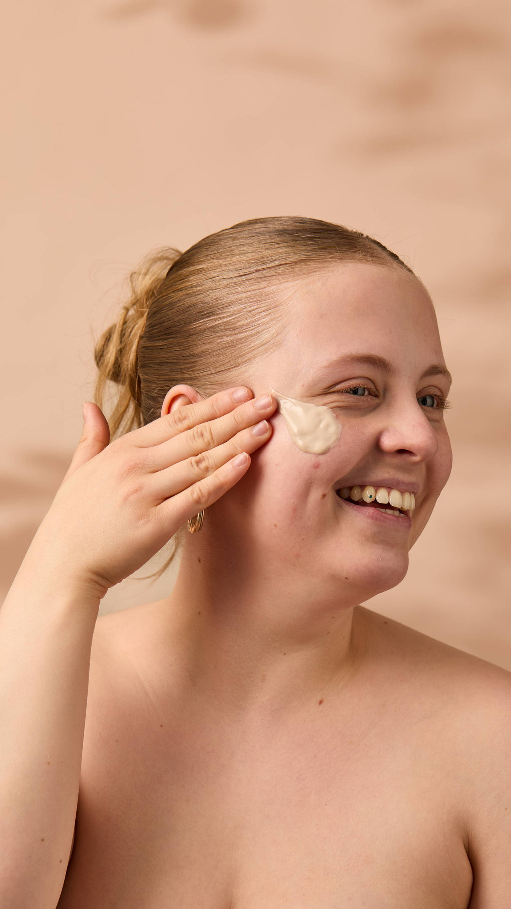 An image of the model's face on a warm, beige background. Their copper hair is tied back, and they have applied the thick Coconut Shie face mask onto their cheek as they smile.