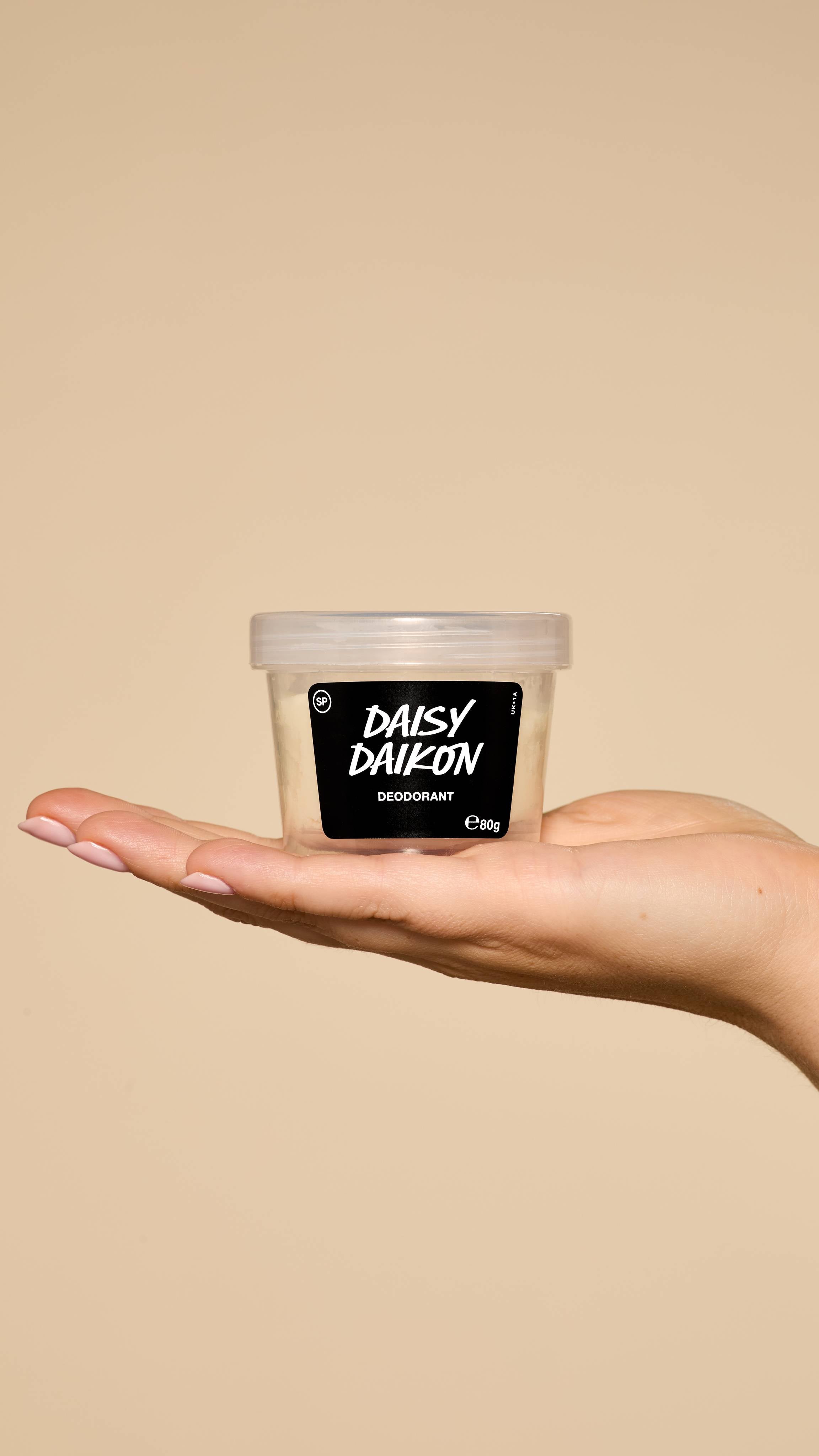 The image shows a warm beige background. A hand reaches in from the right, holding a clear, recycled-plastic pot of Daisy Daikon solid deodorant in a flat palm.