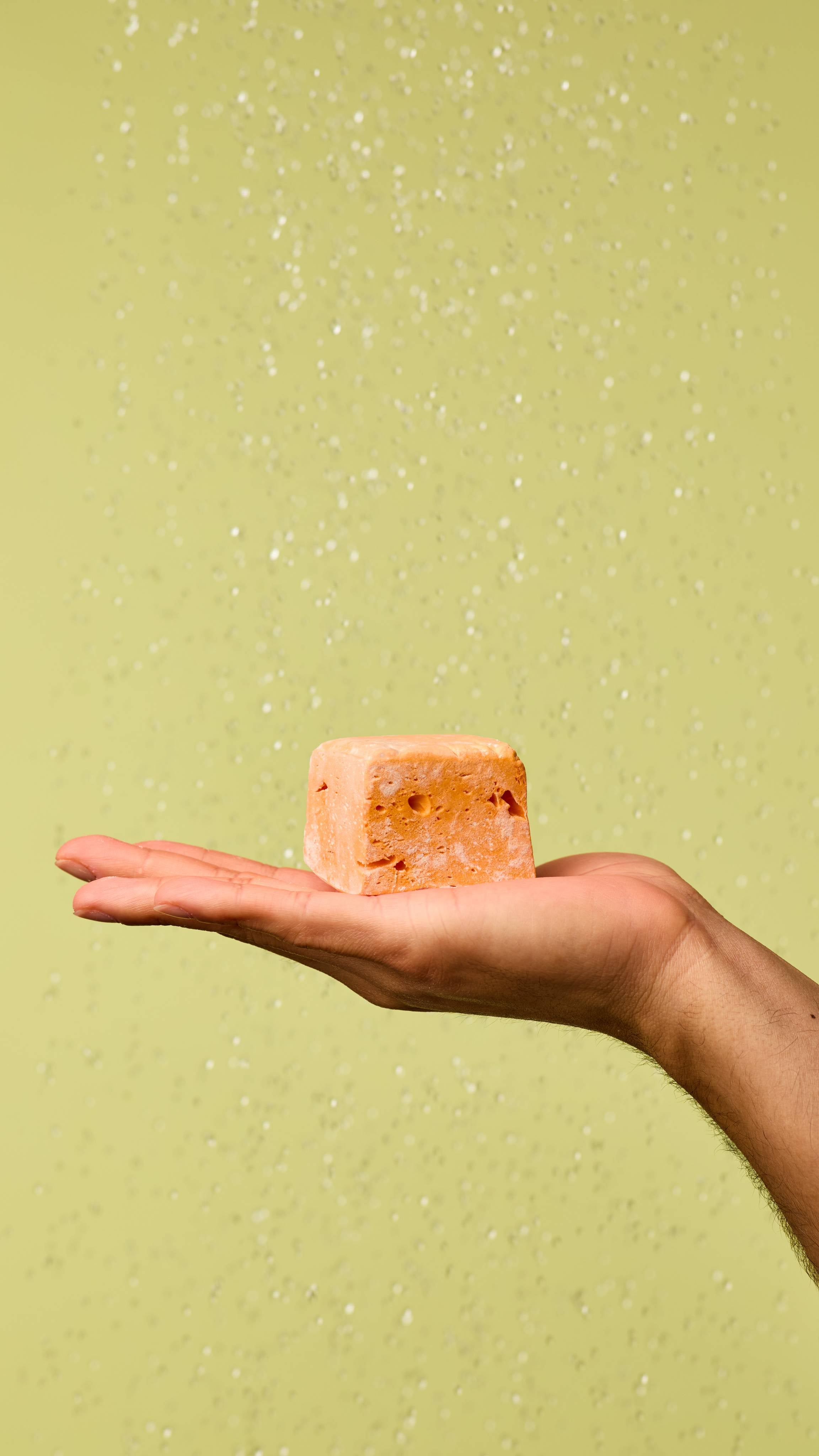 The image shows a warm-toned hand holding a pastel orange Passion Fruit Delight floating soap, resembling a dense marshmallow. Water droplets fall around the hand and soap, against a light green background.