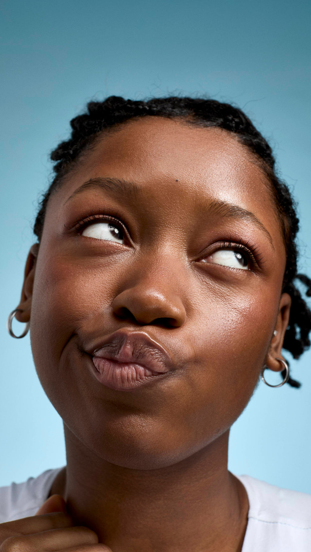 A close-up of a model with braided hair and glowing skin, looking upwards with a playful, pouting expression. They are wearing a white top and small silver hoop earrings against a soft, light-blue background.