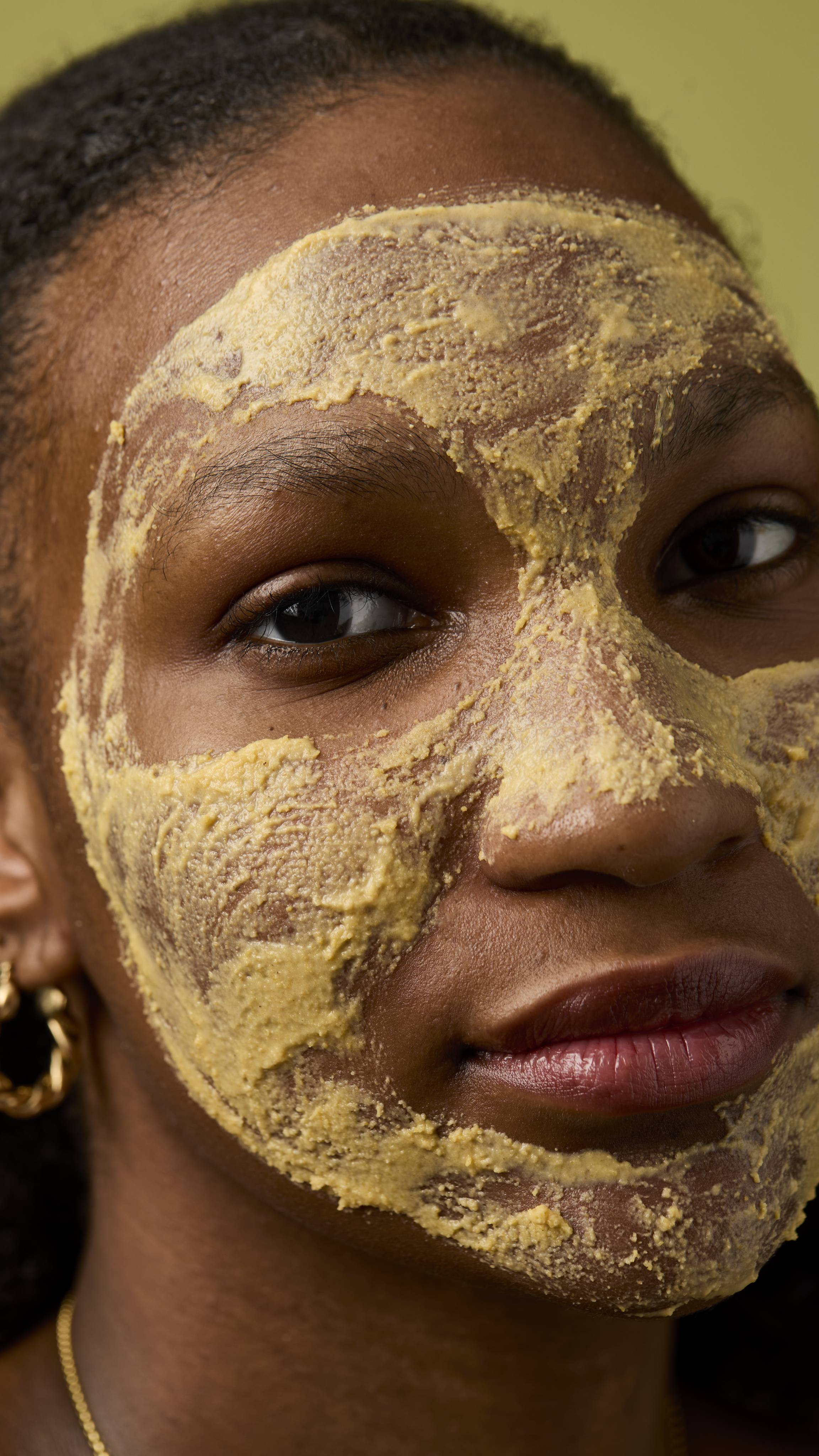 close-up of an individual with the thick, yellow, textured facial cleanser (Let the Good Times Roll) applied fully over their face, covering the forehead, cheeks, and nose. The background is a solid, muted olive-green colour