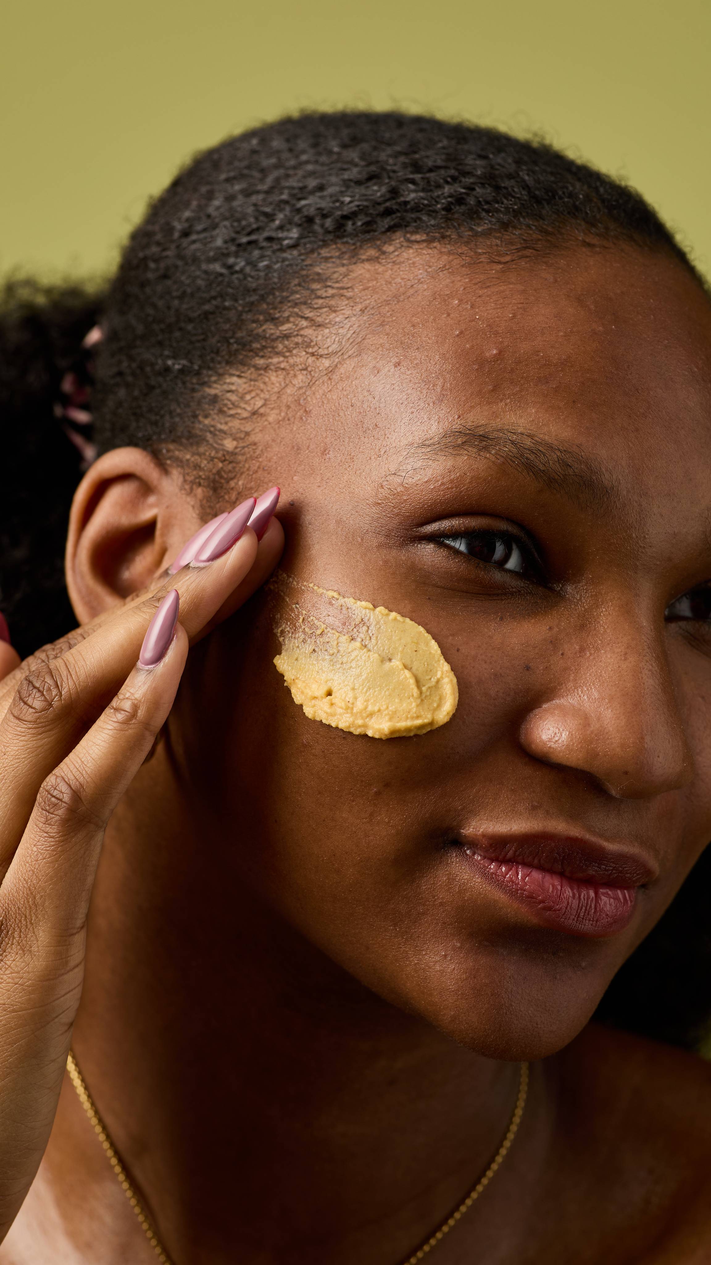 Close-up of an individual applying a dollop of thick, yellow, and textured facial cleanser (Let the Good Times Roll) to their cheek using their fingertips. The background is a solid, muted olive-green color, and the person's hand has manicured nails.