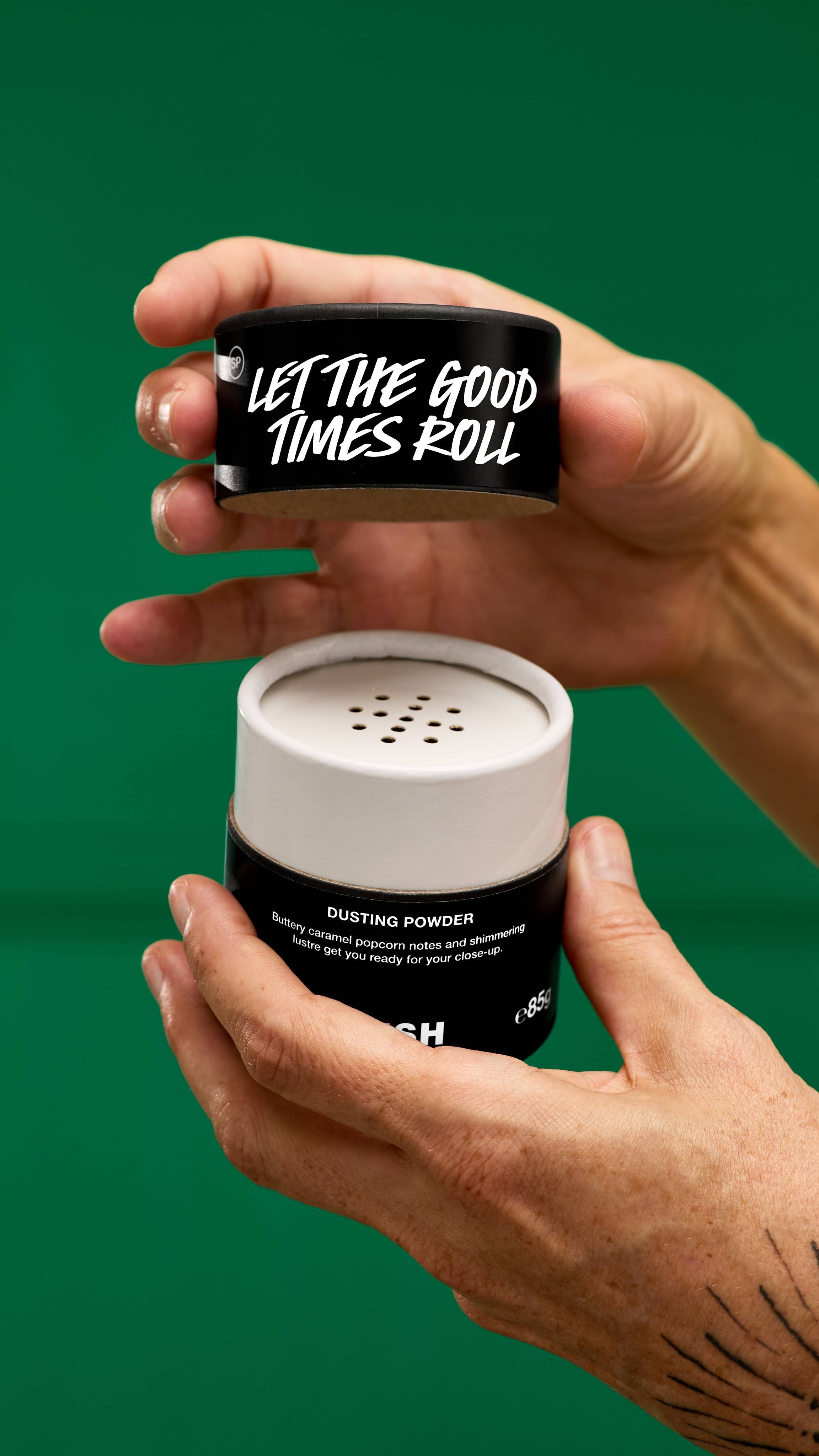 A pair of hands holds open a LUSH Let the Good Times Roll dusting powder container with a black lid and white shaker top, revealing the product inside. The background is a vibrant green.