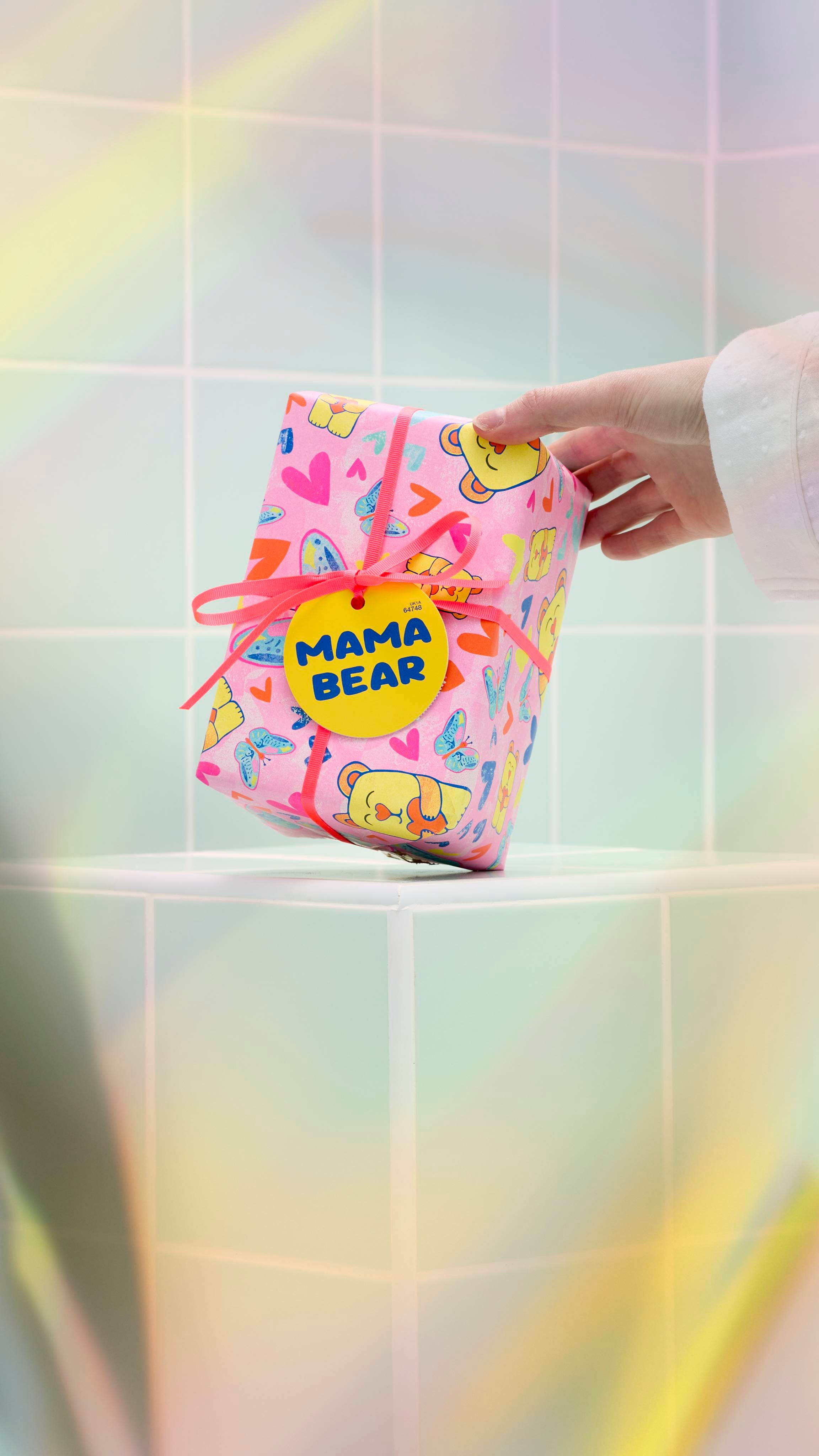 A model's hand reaches out to touch a Lush "Mama Bear" gift, which is wrapped in pink paper decorated with colourful bears, butterflies, and hearts. The gift is tied with a pink ribbon and a yellow tag against a background of light green tiles.
