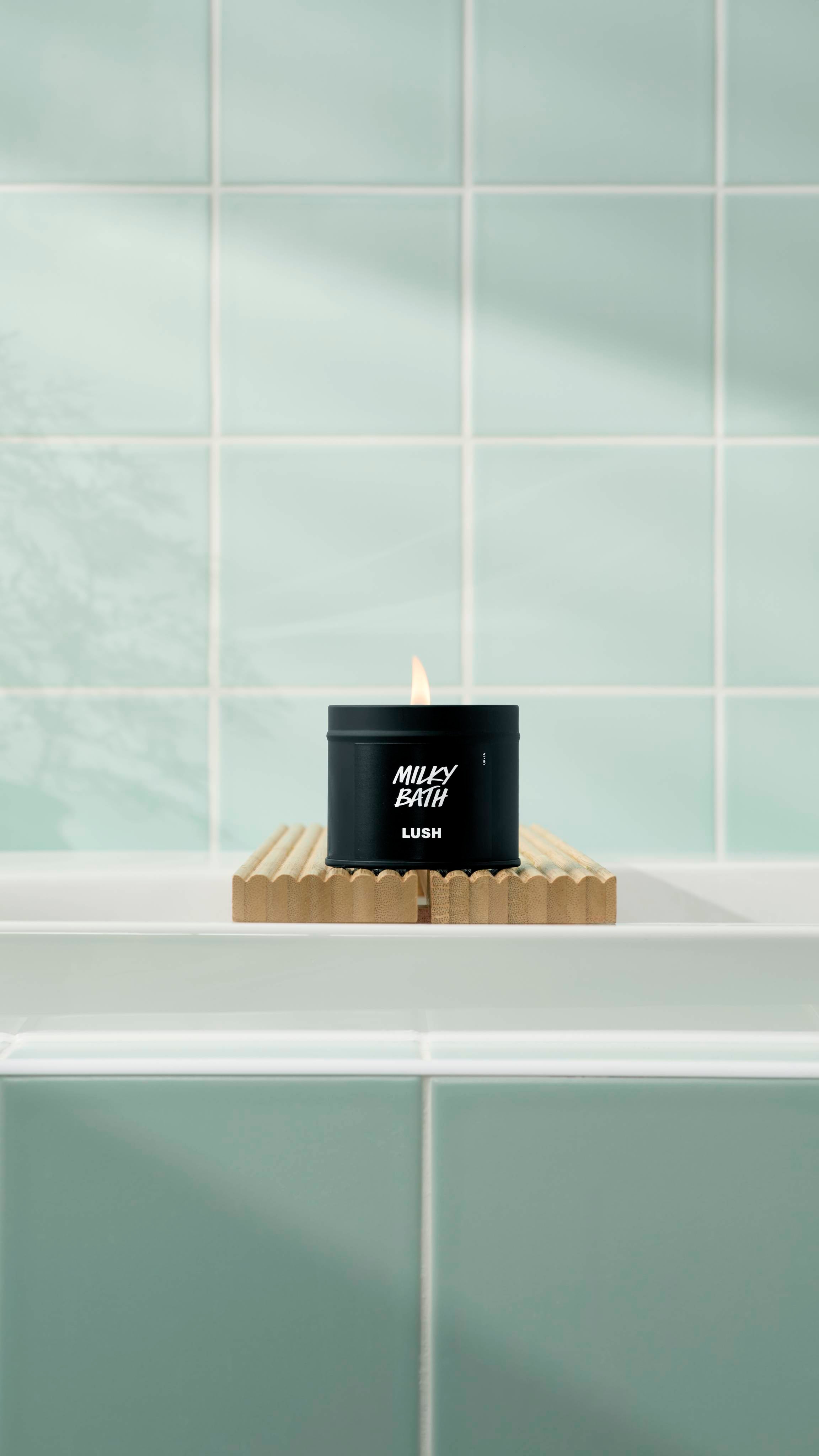 A lit Milky Bath candle in a matte black tin sits on a fluted wooden bath bridge over a white tub. The background features soft mint green square tiles with a gentle shadow of a plant.