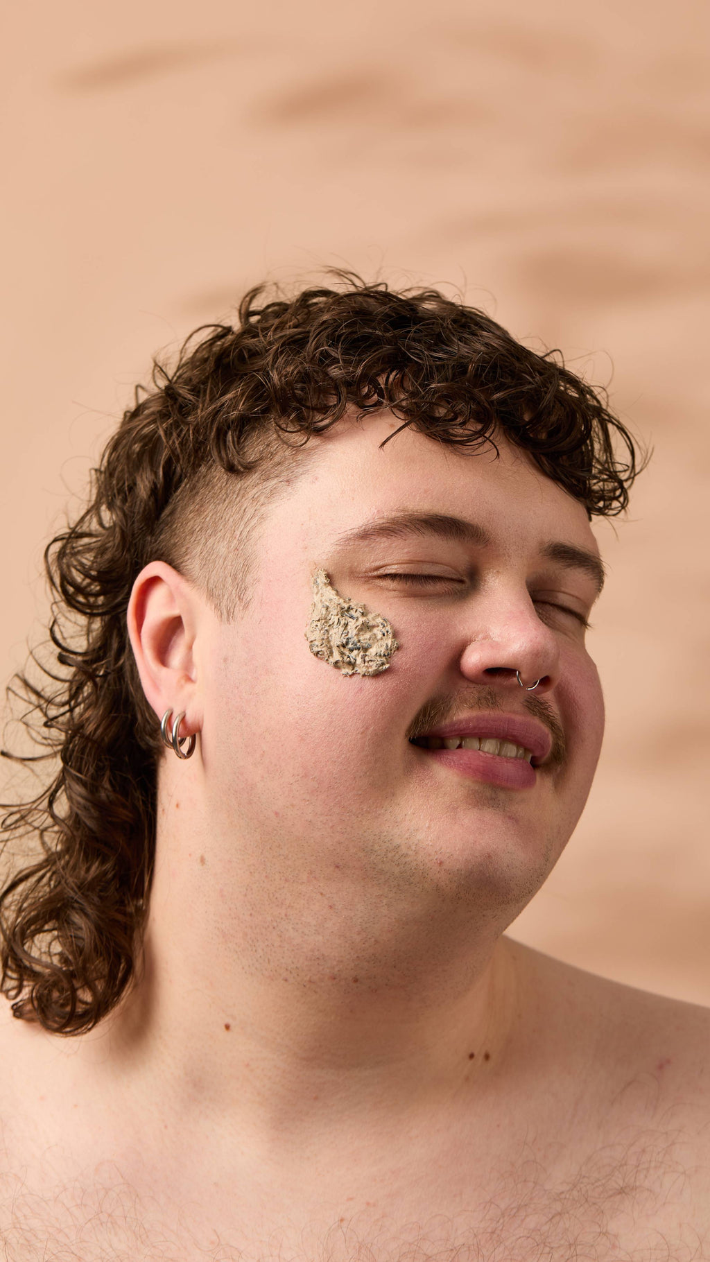 An image of the model's face on a warm, beige background. They have a smudge of the thick, creamy Mineralise Me face mask on their cheek as they smile.