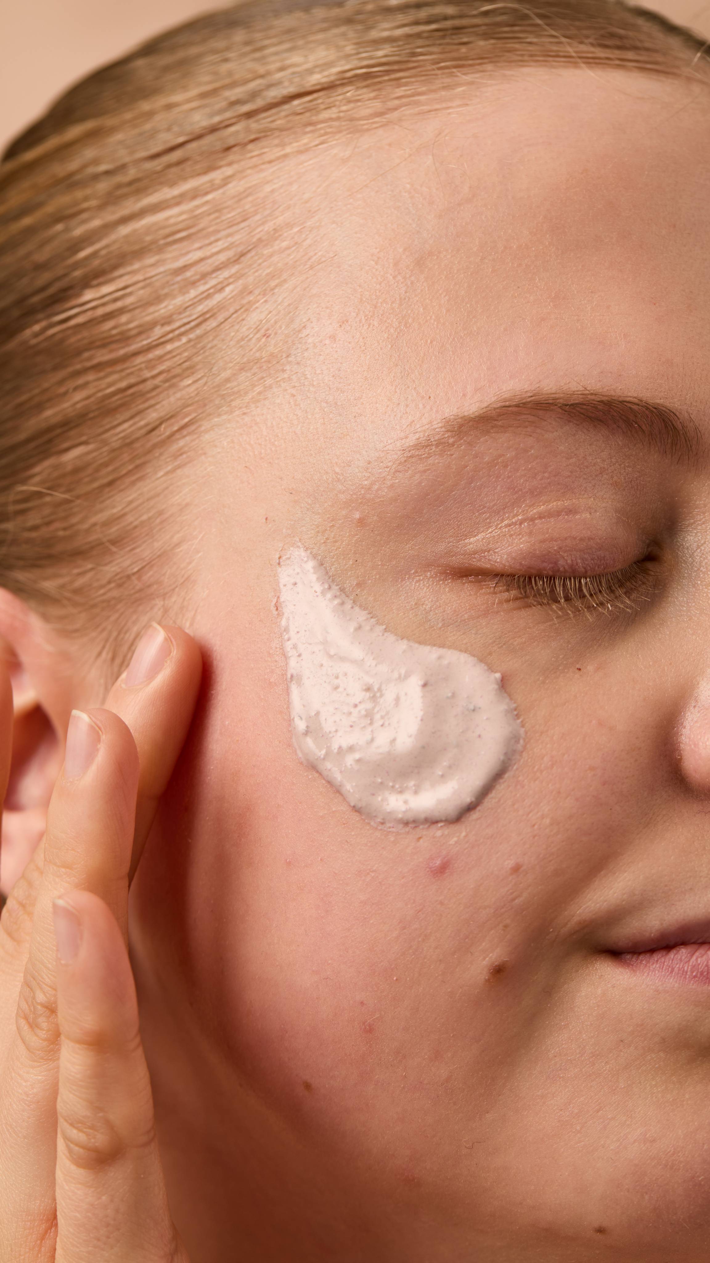 A close-up image of the model's face on a warm, beige background. Their copper hair is tied back, and they have applied the thick Mrs Whippy face mask onto their cheek as their eyes are closed.