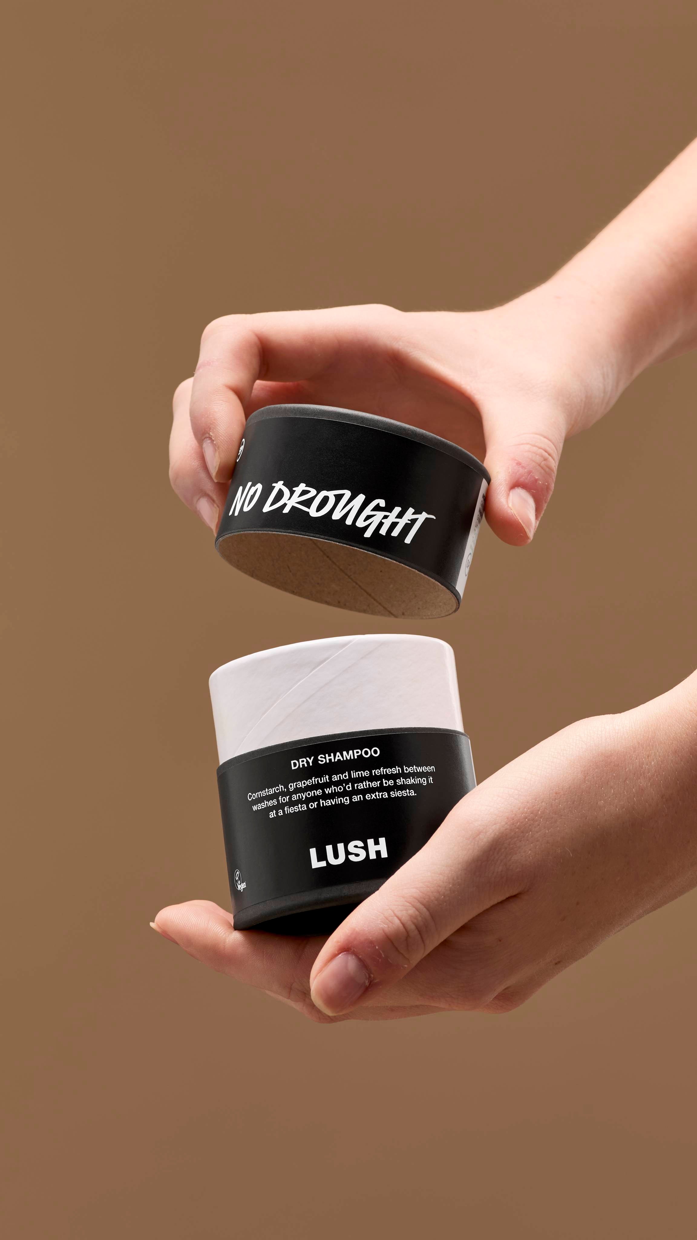 The image shows a pair of hands opening a black and white cardboard tub of 'No Drought' dry shampoo. The lid has white handwritten text, with the tub's black label reading 'DRY SHAMPOO' and 'LUSH', against a solid beige background.