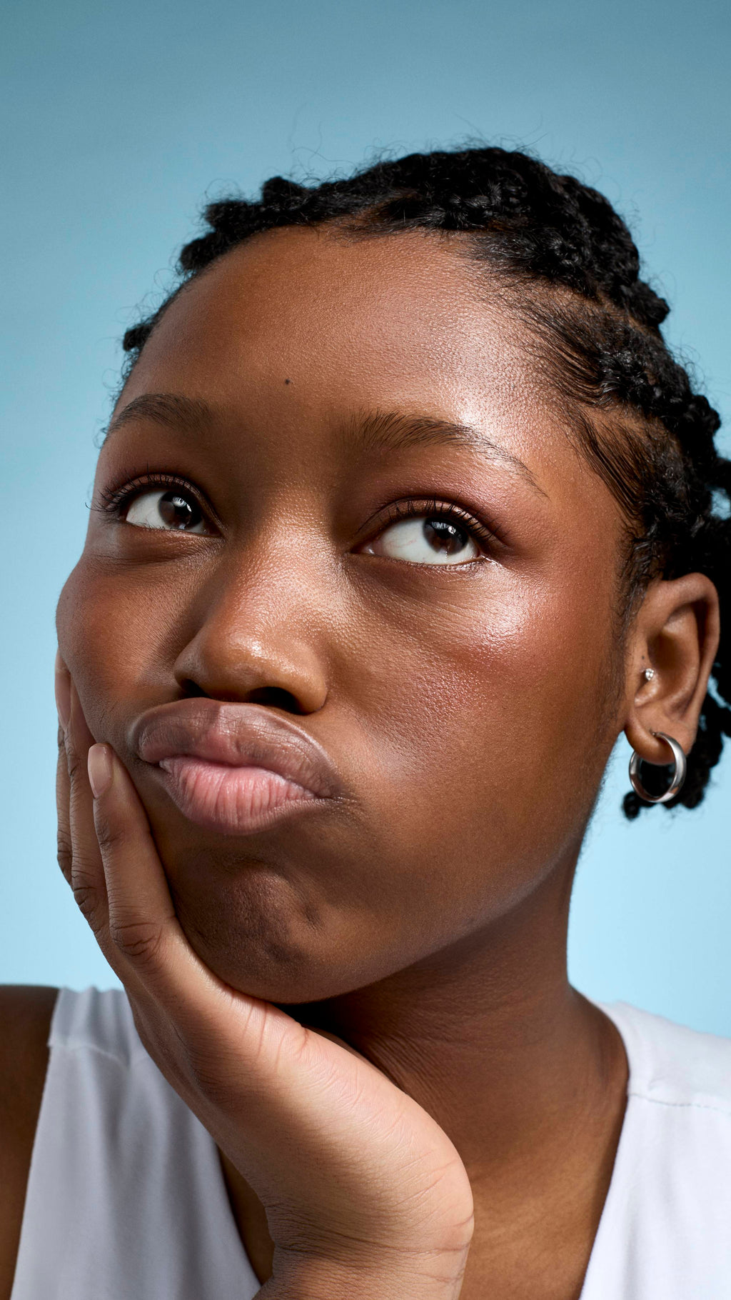 A close-up of a model with deep-toned skin and braided hair, looking upwards with a thoughtful, pouting expression. They are resting their chin in their hand, wearing a white top and small silver hoop earrings against a soft blue background.