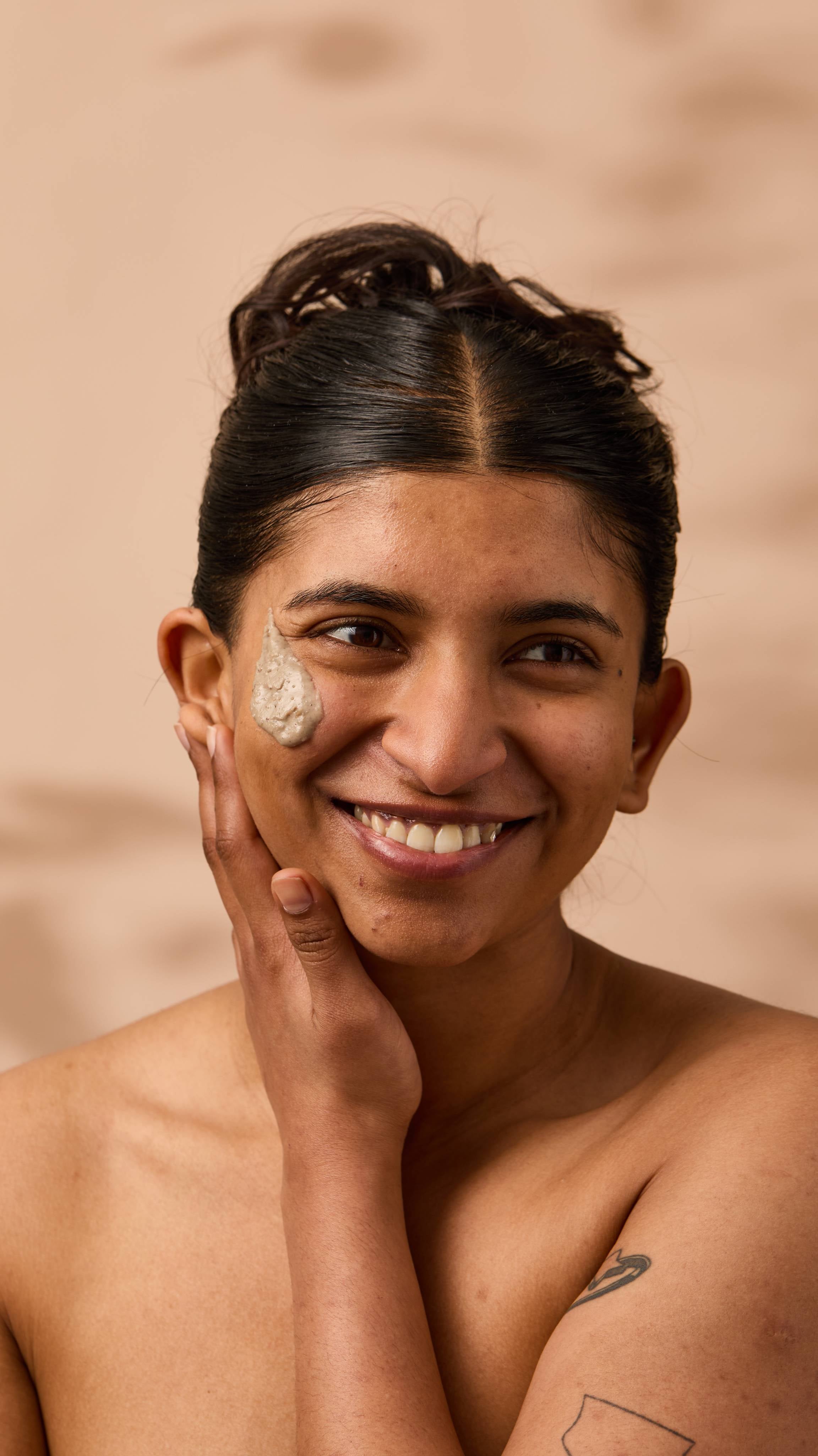 An image of the model's face on a warm, beige background. They have a smudge of the thick, creamy Nutkins face mask on their cheek as they smile.