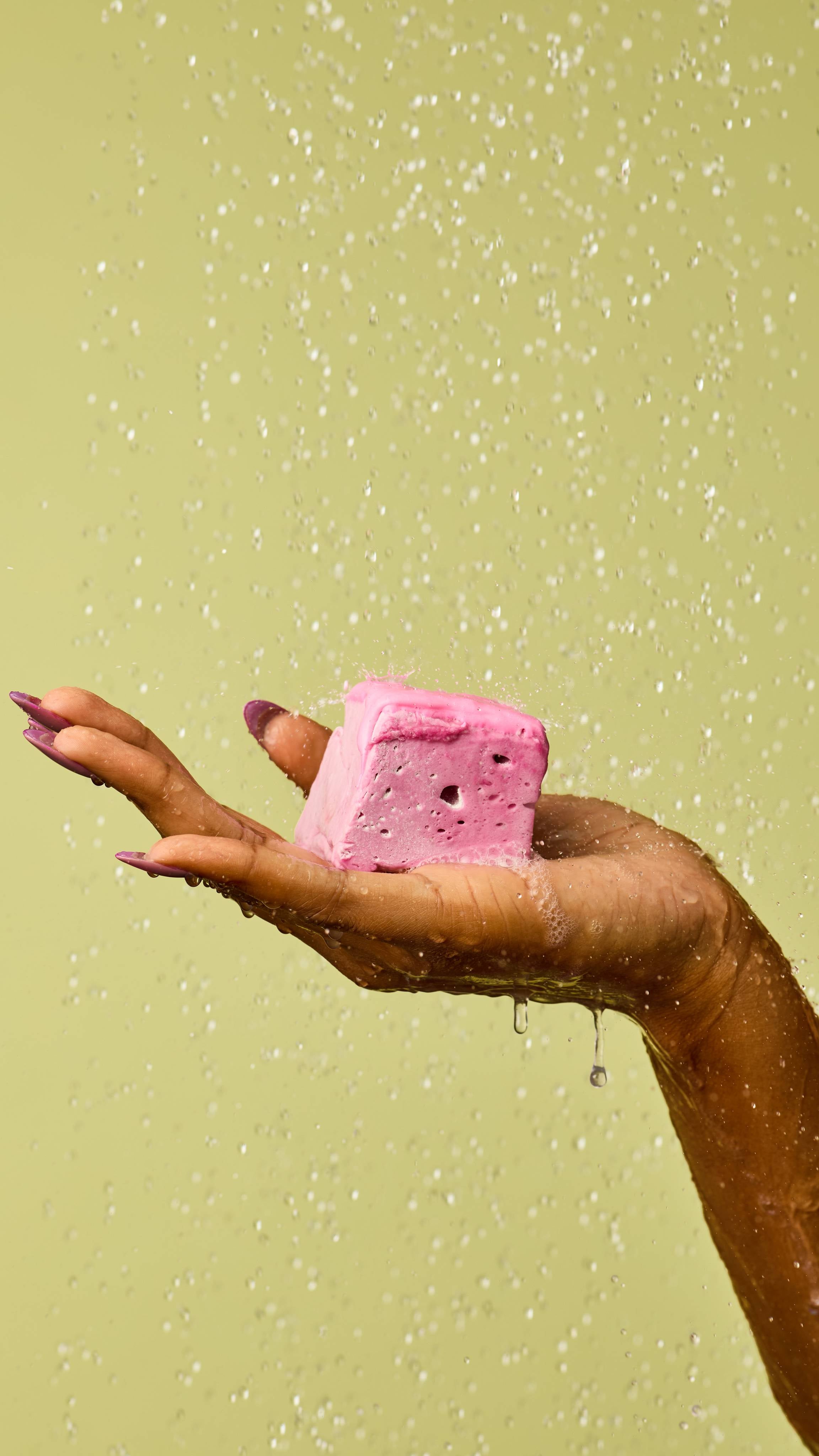 The image shows a hand with light purple nails holding a pastel pink, marshmallow-like Happy Blooming Delight soap cube, topped with a darker pink coating. Water droplets fall around the hand and product against a light green background.