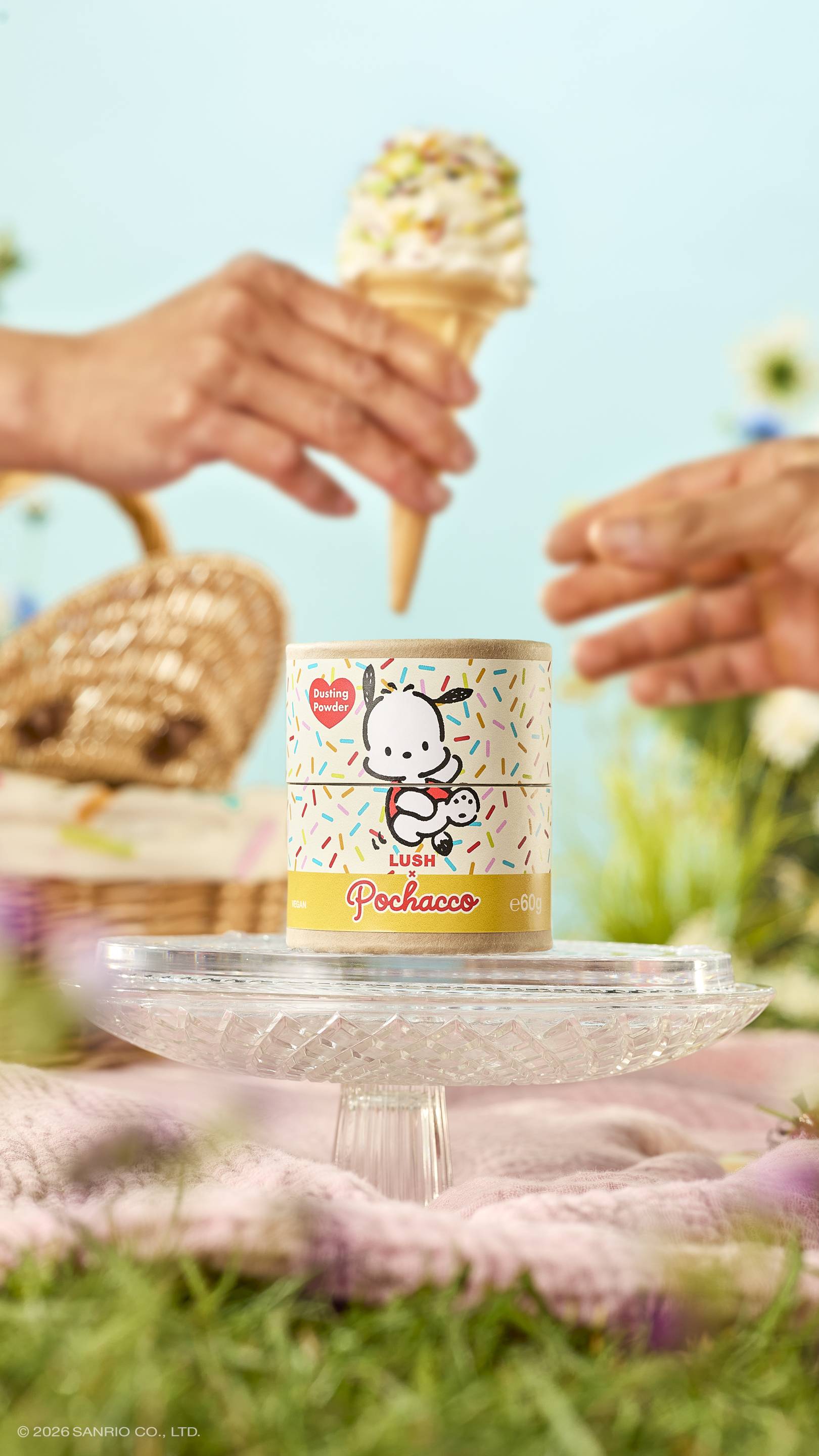 A cardboard tube of Lush x Pochacco dusting powder sits on a glass stand in a meadow. Above it, one hand passes a sprinkle-topped ice cream cone to another. The sunny picnic scene includes a wicker basket and a soft blue sky.