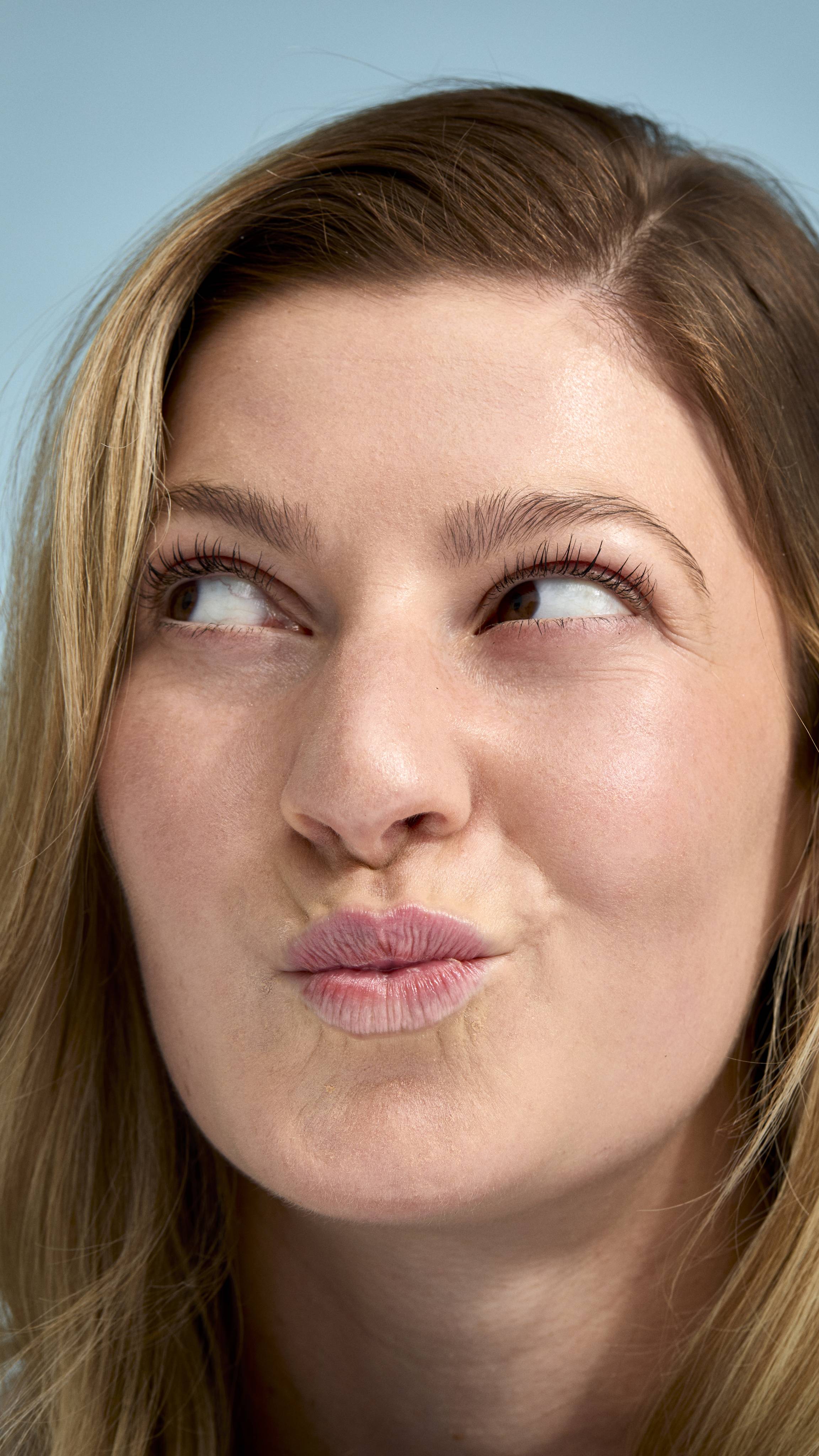 A close-up of a model with fair skin and dark blonde hair looking upwards with a playful, pouting expression. Their face has a natural, glowing finish, set against a soft, light-blue background.
