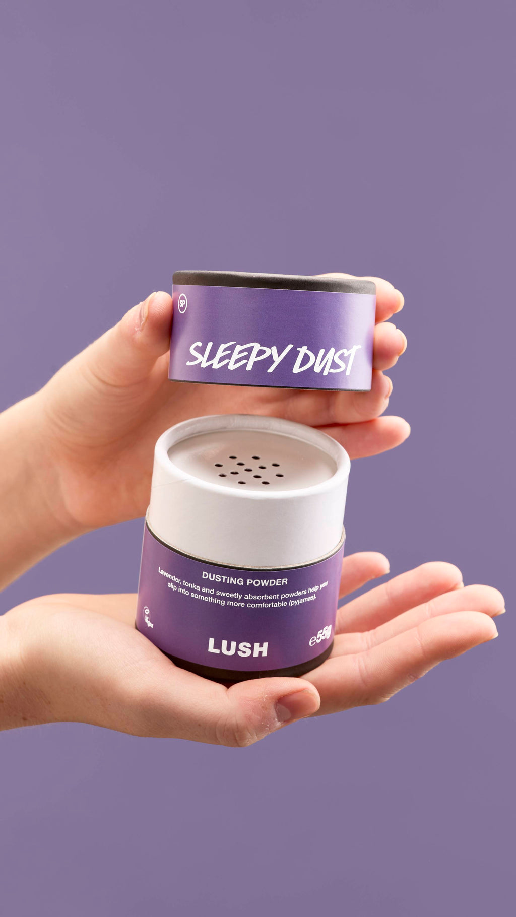 The image shows a pair of hands opening a purple and white cardboard tub of 'Sleepy Dust' dusting powder. The tub has perforated holes on top, with a purple lid with white text, against a solid purple background.