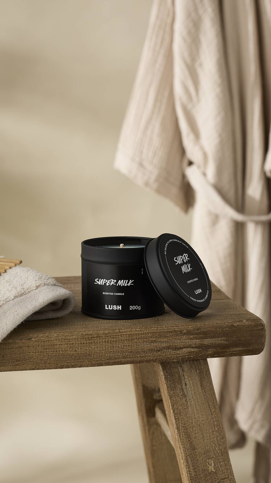 A slightly off-center shot of the black Super Milk Scented Candle tin, sitting open on a rustic, weathered wooden stool. The lid rests leaning on the tin, and a light-coloured towel and a beige bathrobe hang in the soft-focus background.