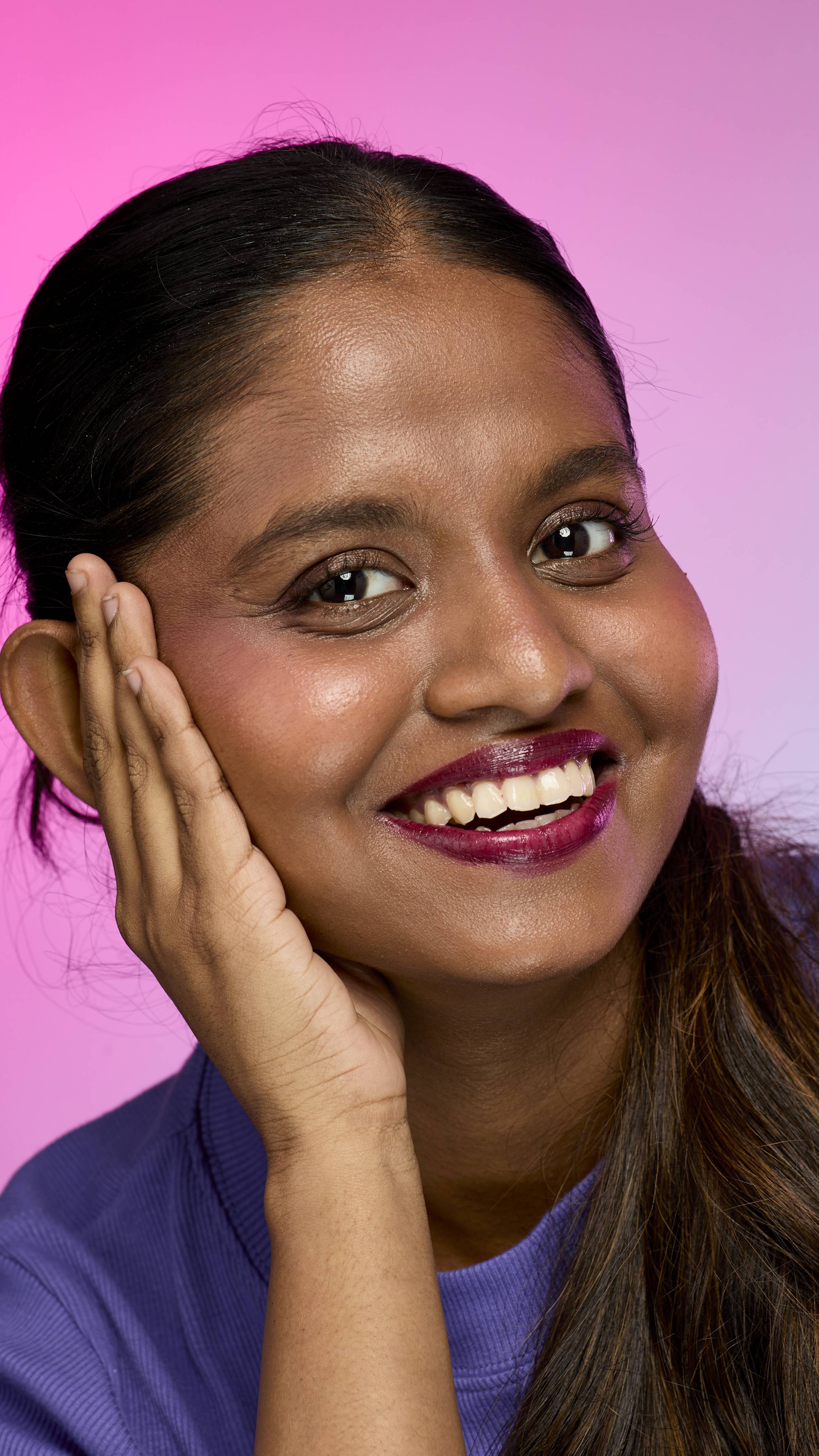 A smiling person with dark hair and skin, wearing a purple top, is shown from the chest up. They are wearing the Lush, deep plum Tartlet lip cream and have one hand touching the side of their face. The background is a pink and purple gradient.