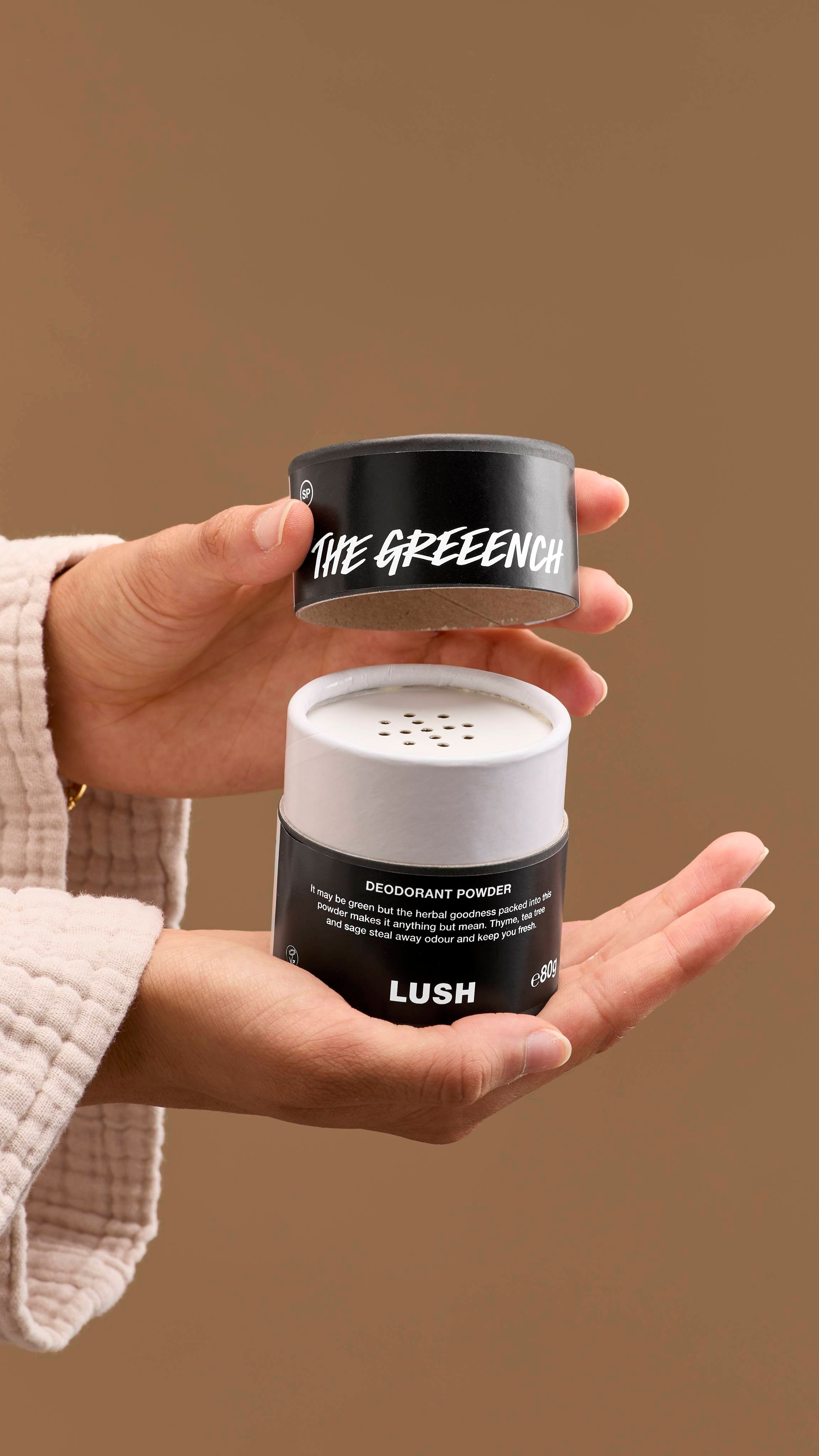 The image shows a pair of hands opening a white and black, cardboard tub of 'The Greench' deodorant powder. The tub has perforated holes on top, with a black lid with white text 'THE GREENCH', against a solid beige background.