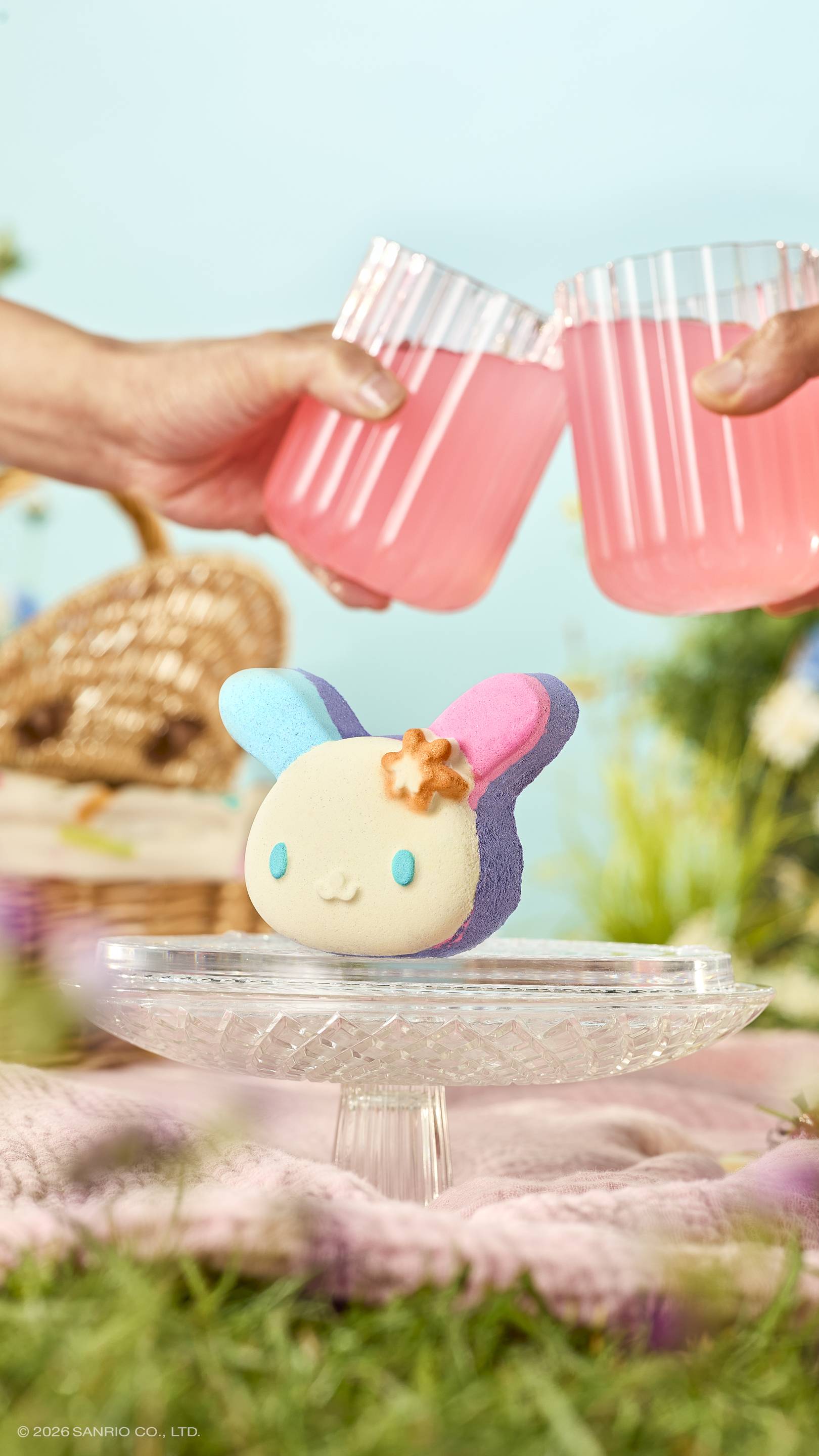The bunny-shaped "U.SA.HA.NA" bath bomb with blue and pink ears sits on a glass cake stand in a meadow. Above it, two hands clink glasses of pink juice in a toast. The sunny picnic scene includes a wicker basket and a soft blue sky.