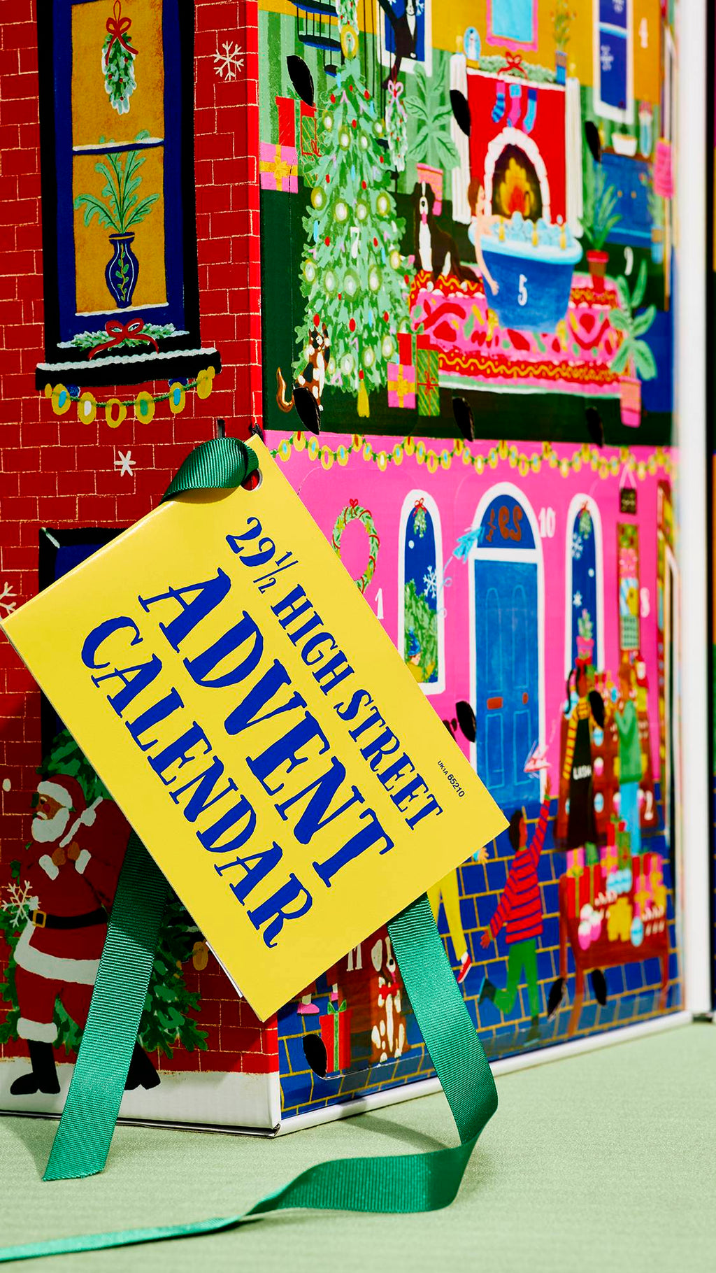 A close-up of a house-themed advent calendar featuring festive illustrated scenes, adorned with a yellow tag tied by a green ribbon that reads "29 ½ High Street Advent Calendar" in blue lettering.