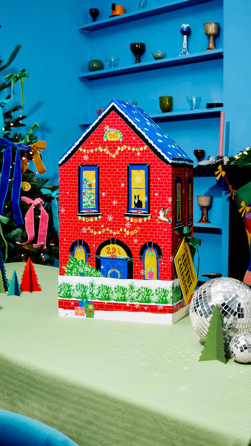 A red brick house-shaped advent calendar with a blue snow-dusted roof stands on a green table, decorated with Christmas lights, a black cat in the window, and a yellow tag attached, set beside silver disco balls.