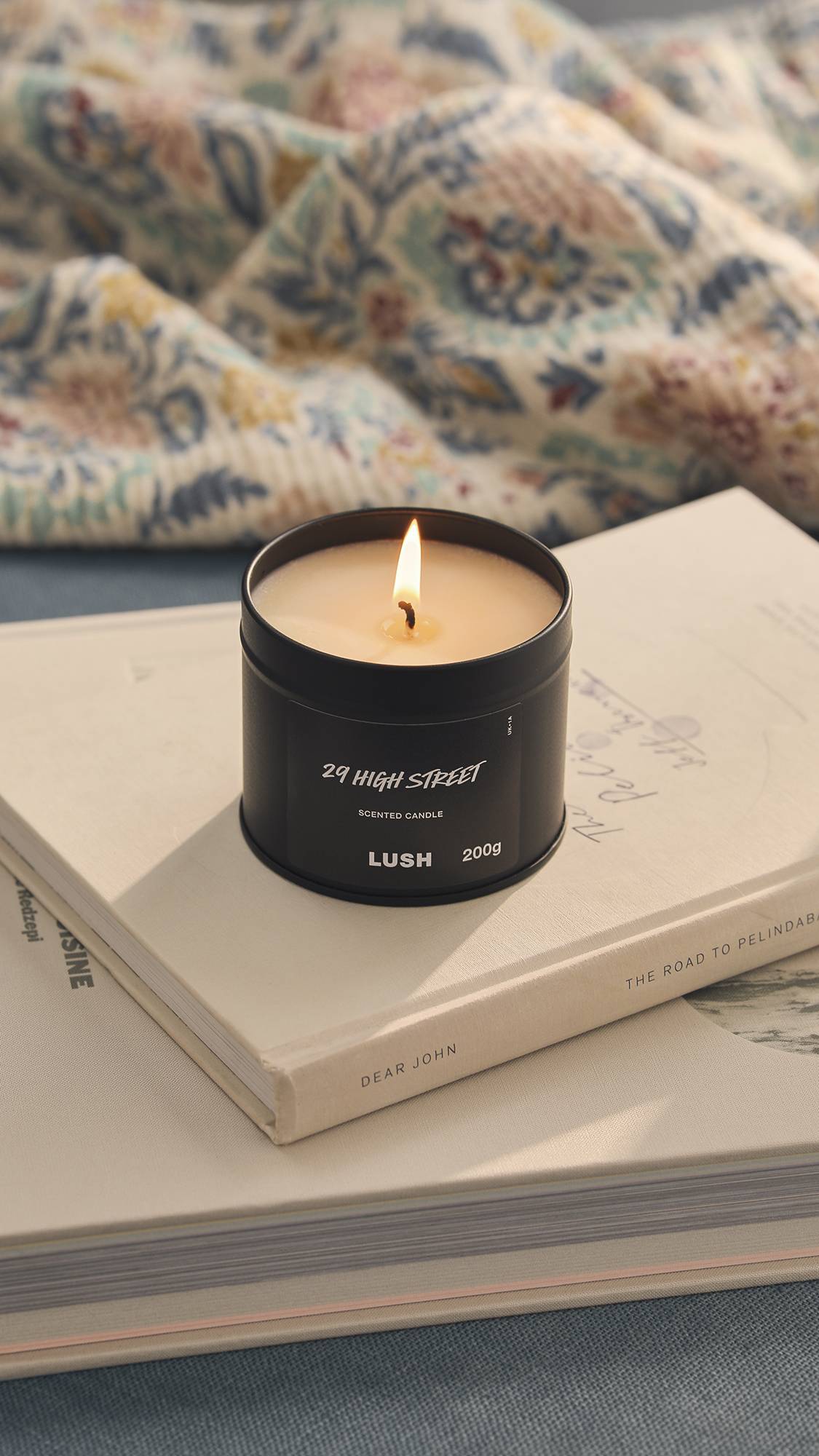 The image shows a blurred background of a patterned blanket. The foreground shows the lit black tin 29 High Street candle sitting on top of a small pile of books.