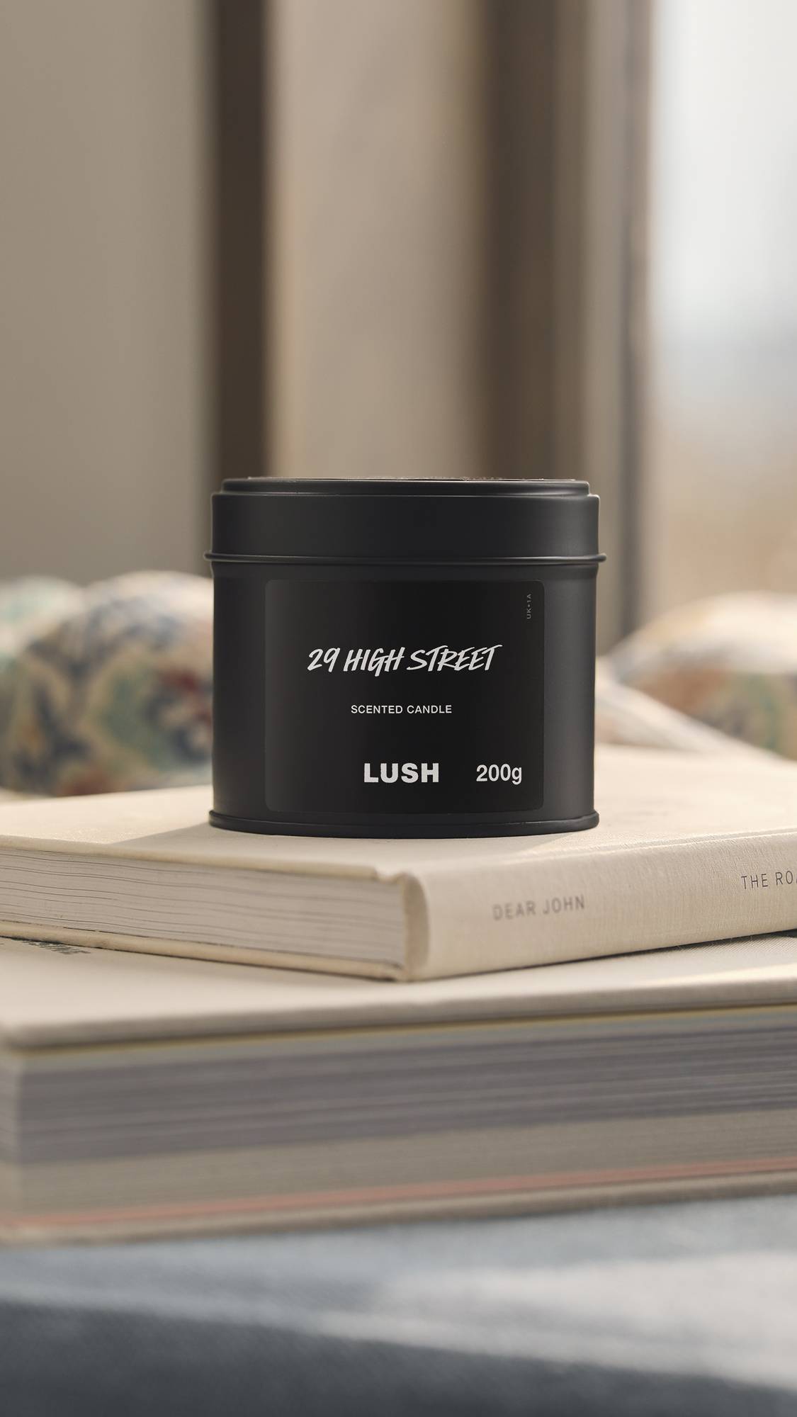 The image shows a natural, bright bedroom background. The foreground shows the black tin 29 High Street candle sitting on top of a small pile of books.