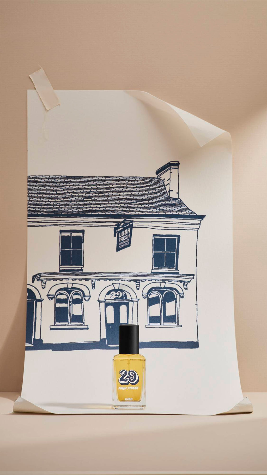 A glass bottle of Lush '29 High Street' perfume stands in front of a large cream poster. The poster features a navy-blue line drawing of the original Lush shop facade. The scene is set against a neutral beige background with soft lighting.