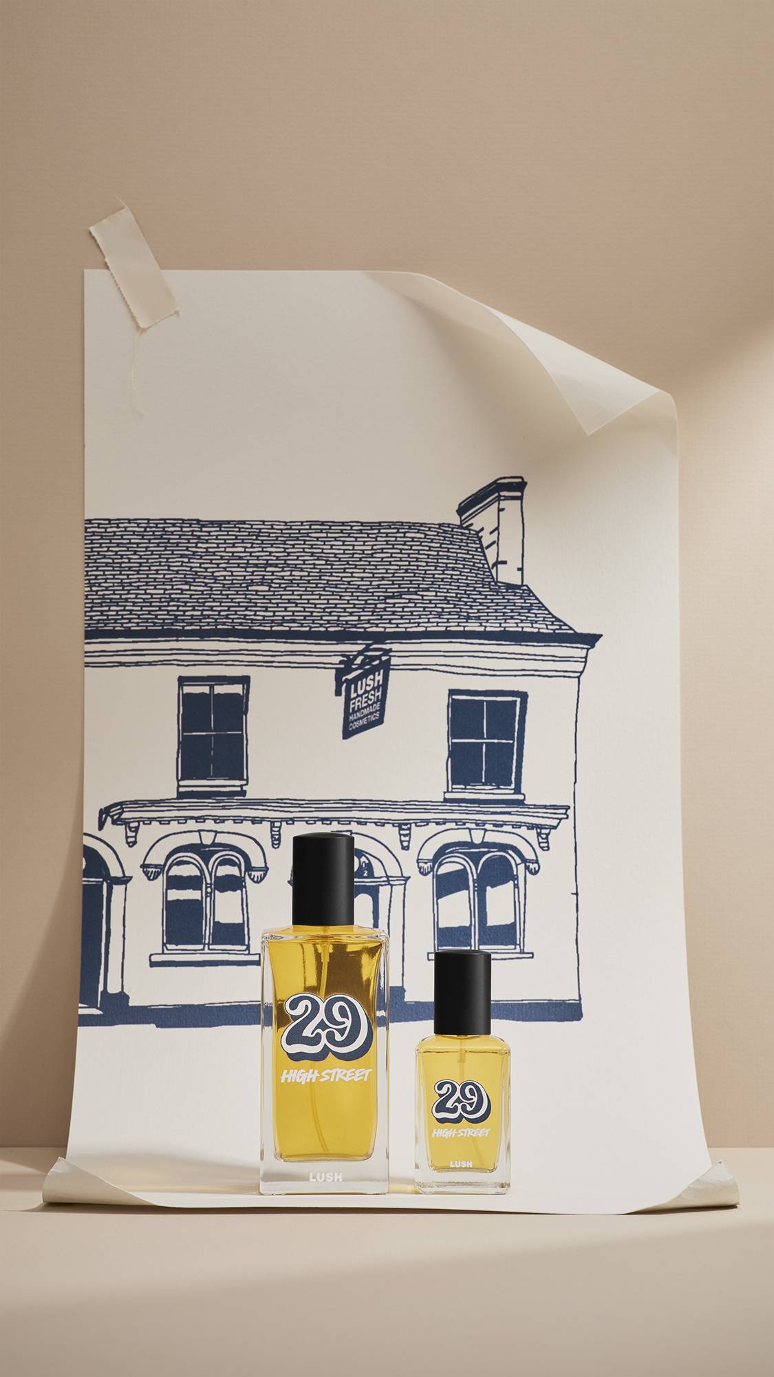 Two glass bottles of Lush '29 High Street' perfume, 30ml and 100ml, stand in front of a large cream poster. The poster features a navy-blue line drawing of the original Lush shop. The scene is set against a neutral beige background.