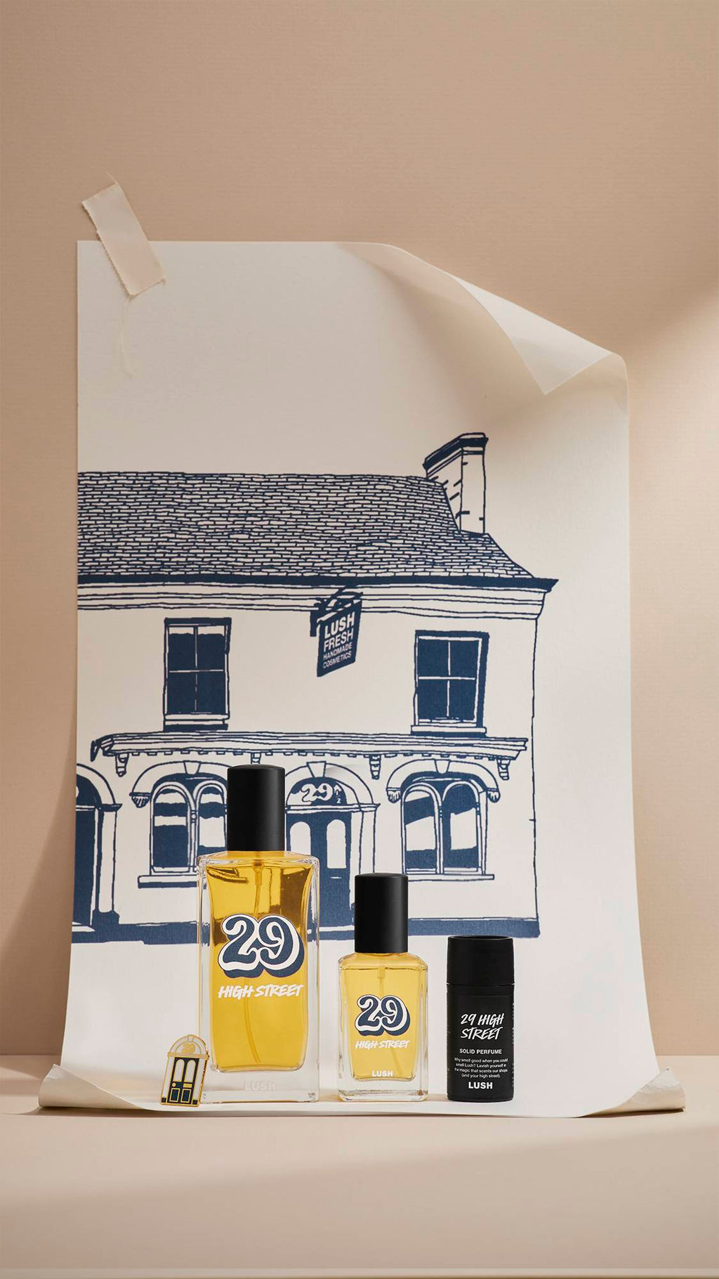 Four products from the Lush '29 High Street' range—two glass perfume bottles, a metal pin badge and a solid perfume tin—stand in front of a navy-blue line drawing of the original Lush shop on a large poster.
