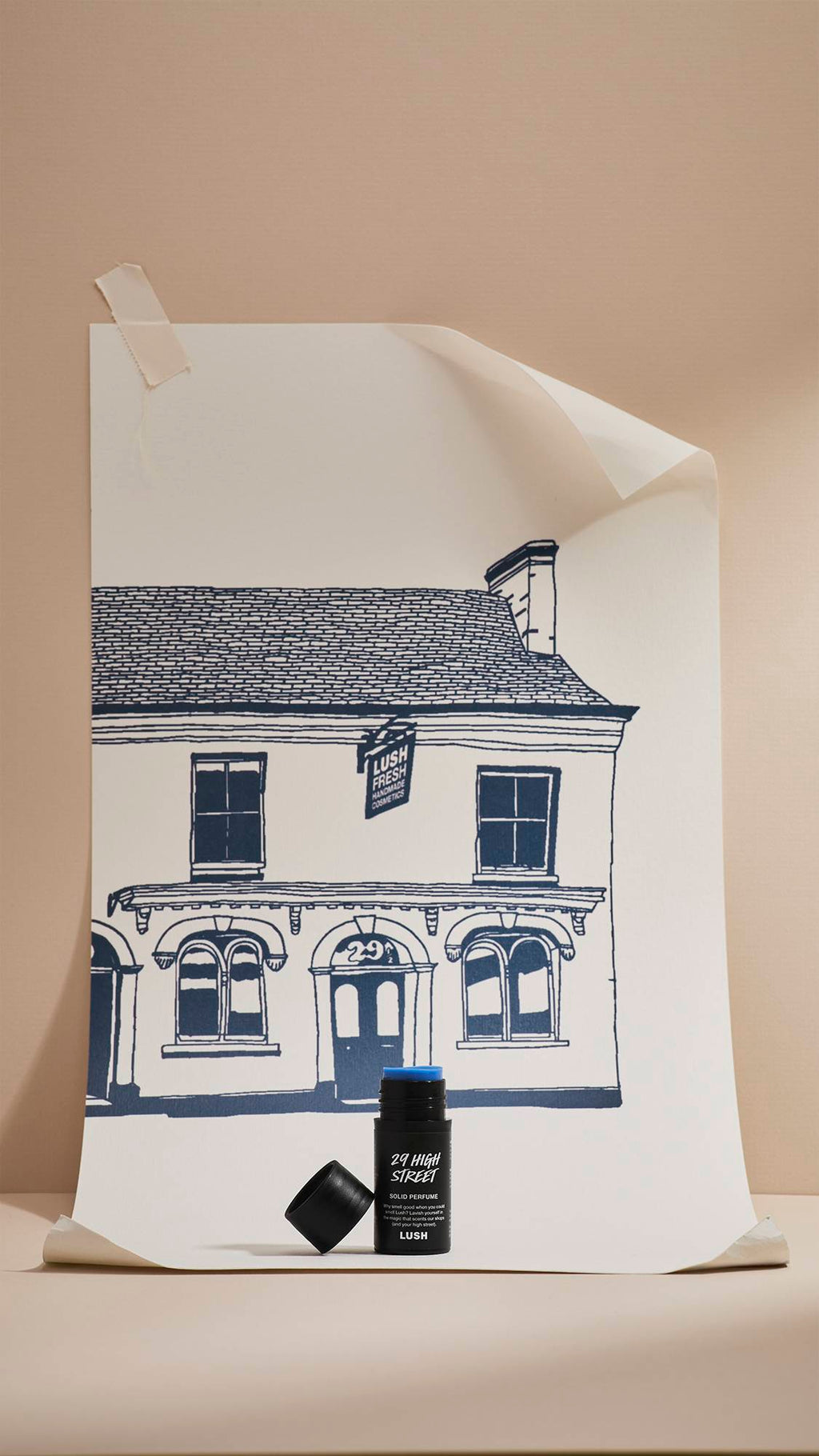 An open black tube of Lush '29 High Street' solid perfume, showing a blue solid balm inside, stands in front of a cream poster. The poster features a navy-blue line drawing of a shop facade, set against a neutral beige background.