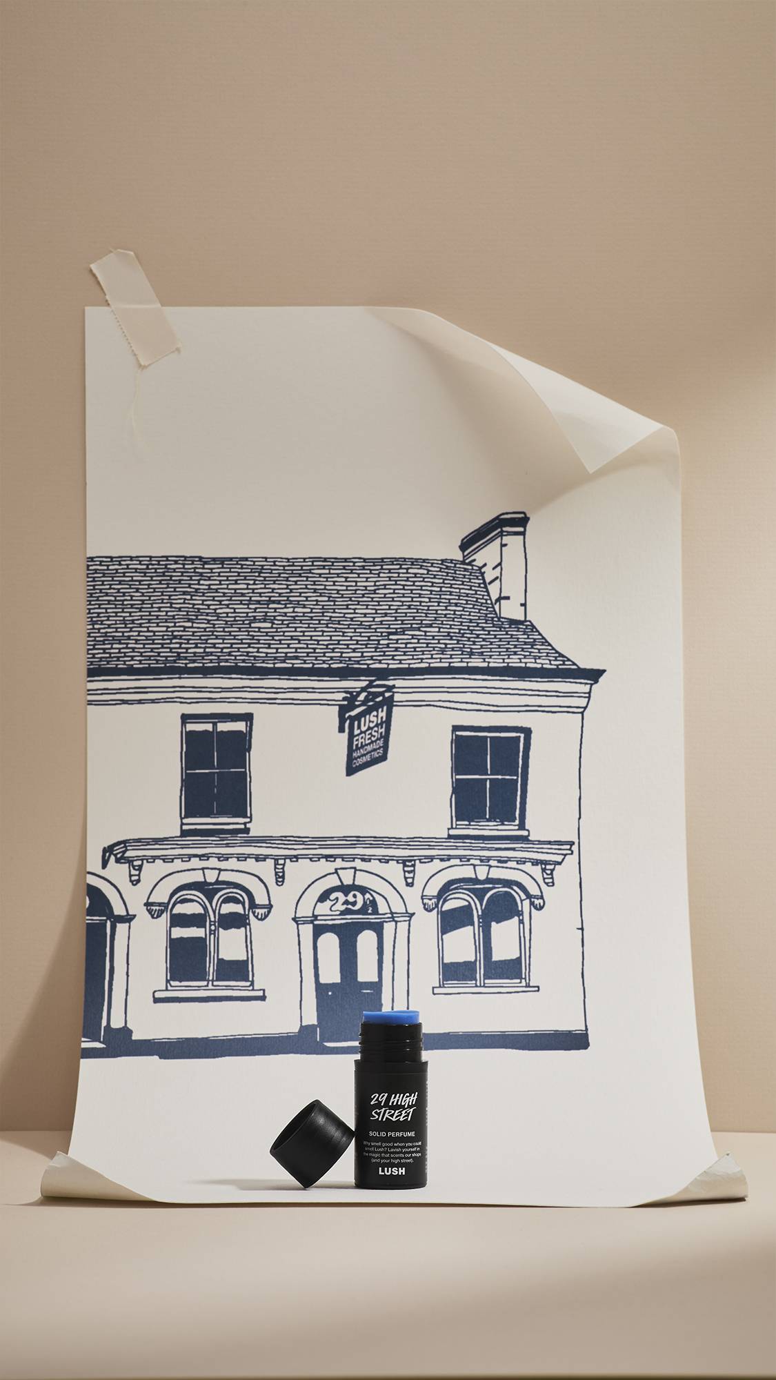An open black tube of Lush '29 High Street' solid perfume, showing a blue solid balm inside, stands in front of a cream poster. The poster features a navy-blue line drawing of a shop facade, set against a neutral beige background.
