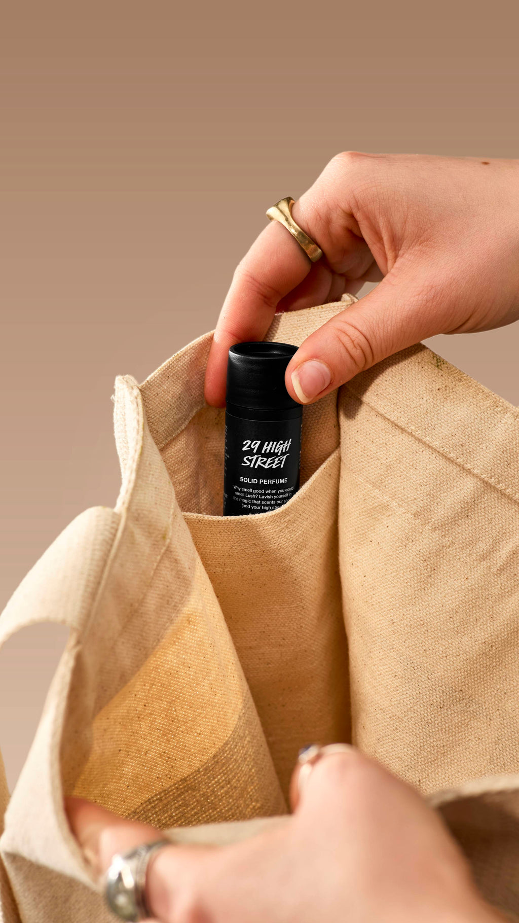 A close-up shot shows a hand placing a small, black Lush '29 High Street' solid perfume tube into the inner pocket of a cream canvas tote bag. The person is wearing several rings, and the image is set against a neutral beige background.