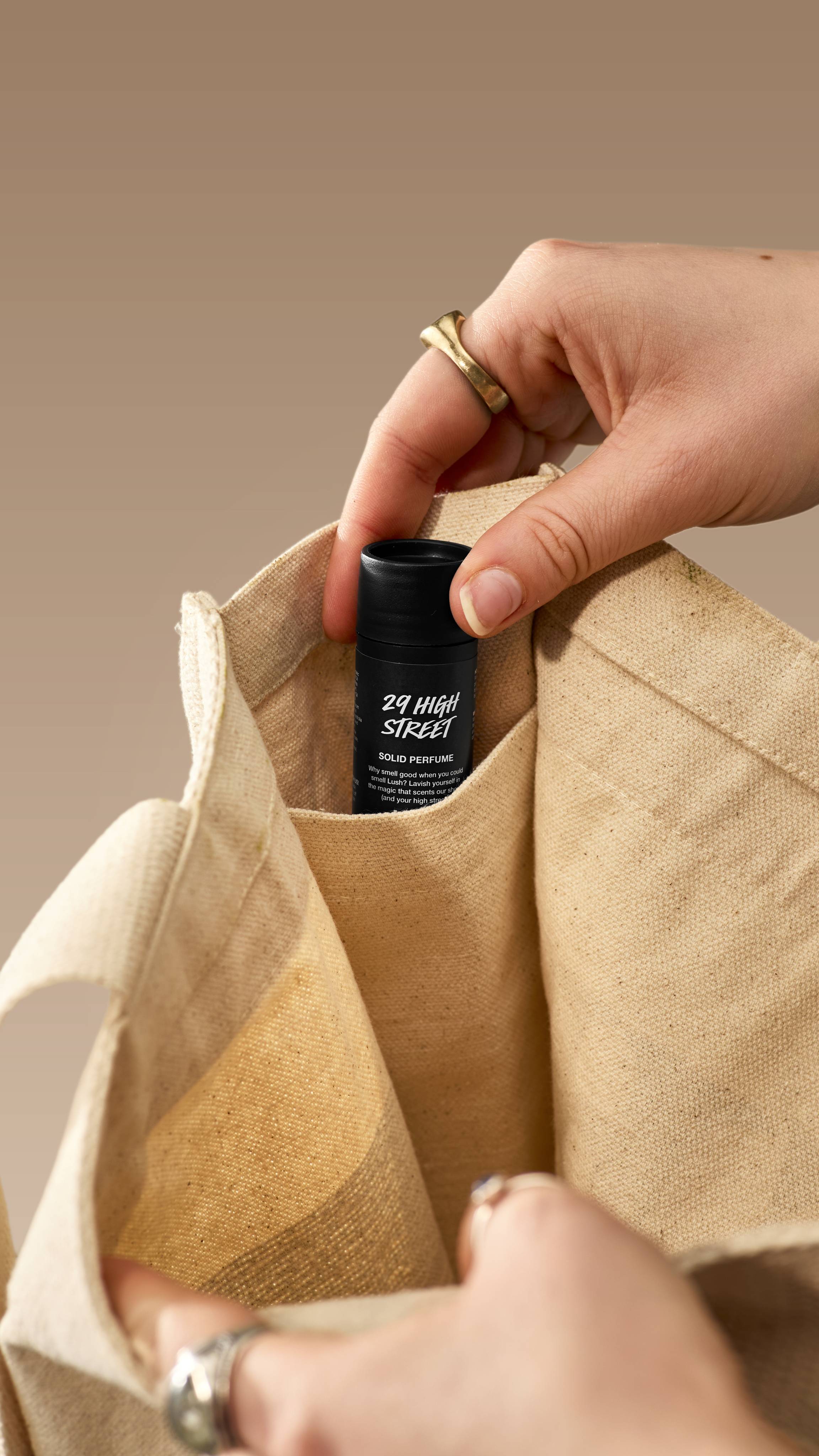 A close-up shot shows a hand placing a small, black Lush '29 High Street' solid perfume tube into the inner pocket of a cream canvas tote bag. The person is wearing several rings, and the image is set against a neutral beige background.