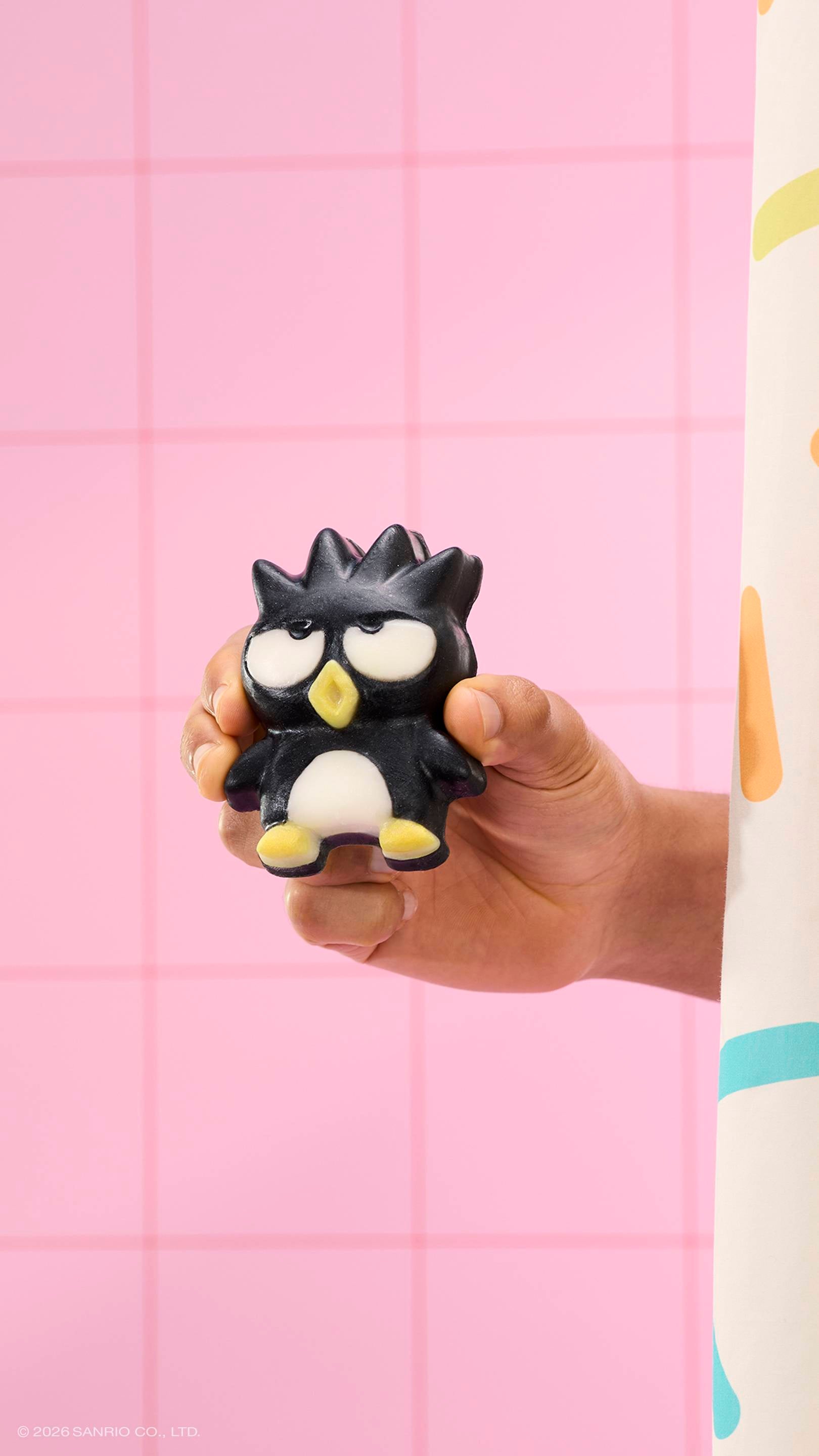 A hand holds a black penguin-shaped soap with spiky hair and a grumpy expression against a pink tiled wall. A sprinkle-patterned shower curtain is visible on the right. The text '© 2026 SANRIO CO., LTD.' is at the bottom.