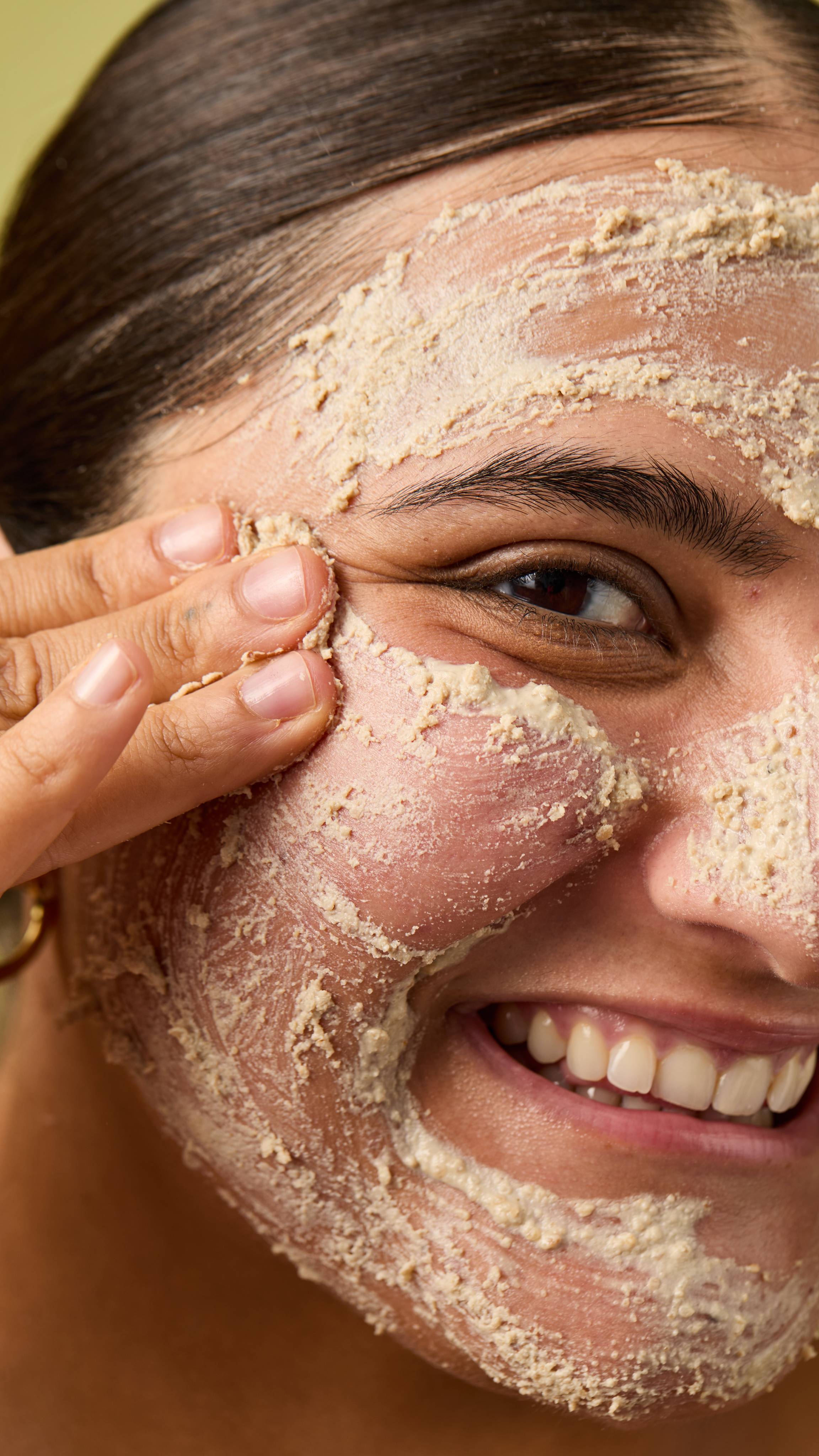 A close-up image of half of the model's face, which is coated in a layer of the textured Biomic facial cleanser as they smile at the camera, on an olive-green background.