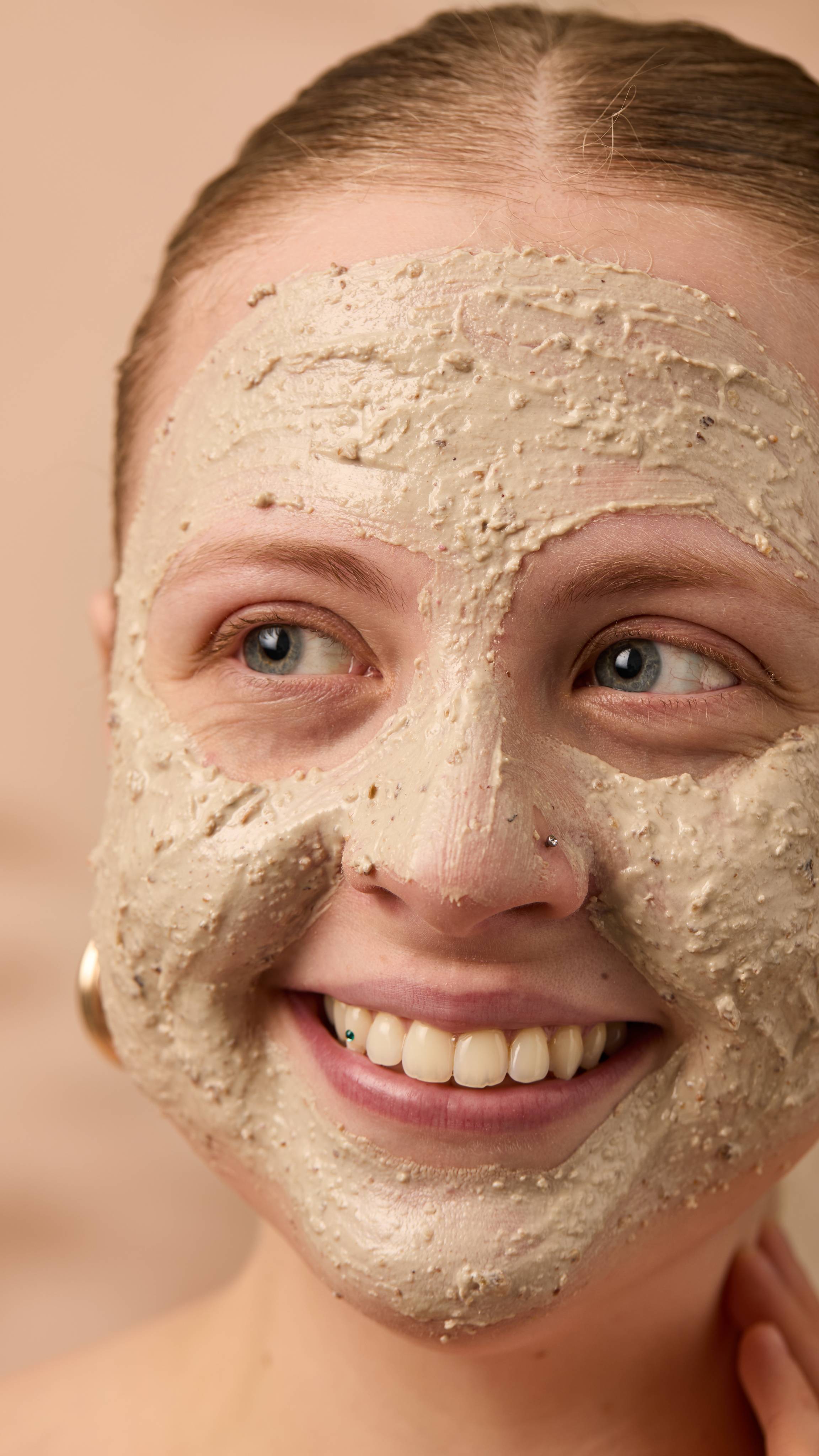 A close-up image of the model's face on a warm, beige background. Their copper hair is tied back, and they have applied the textured Bring Us Some Figgy Pudding face mask all over their face as they smile.